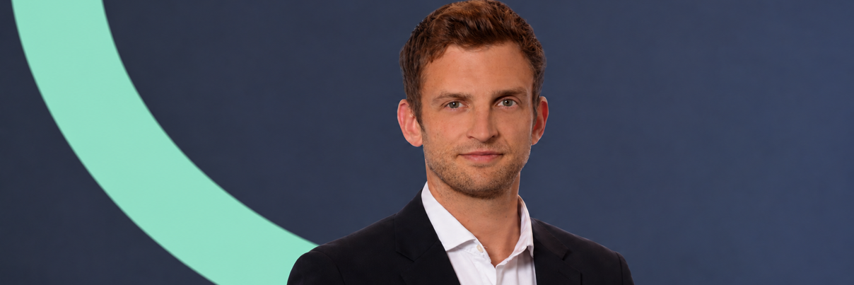 A man with short brown hair wearing a white dress shirt and dark blazer stands against a dark blue and light green background.