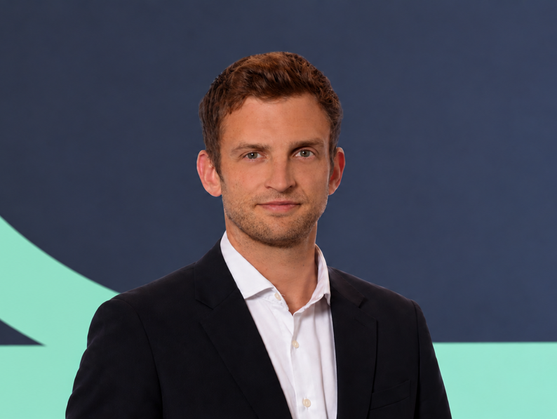 A man with short brown hair wearing a white dress shirt and dark blazer stands against a dark blue and light green background.