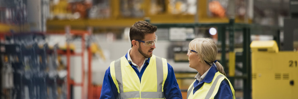 Two workers in high-visibility vests and safety glasses are talking in a large industrial facility. One holds a tablet while listening as the other gestures, with machinery and equipment visible in the background.
