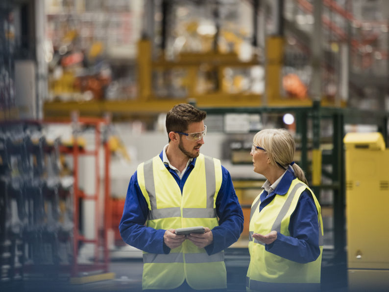 Two workers in high-visibility vests and safety glasses are talking in a large industrial facility. One holds a tablet while listening as the other gestures, with machinery and equipment visible in the background.
