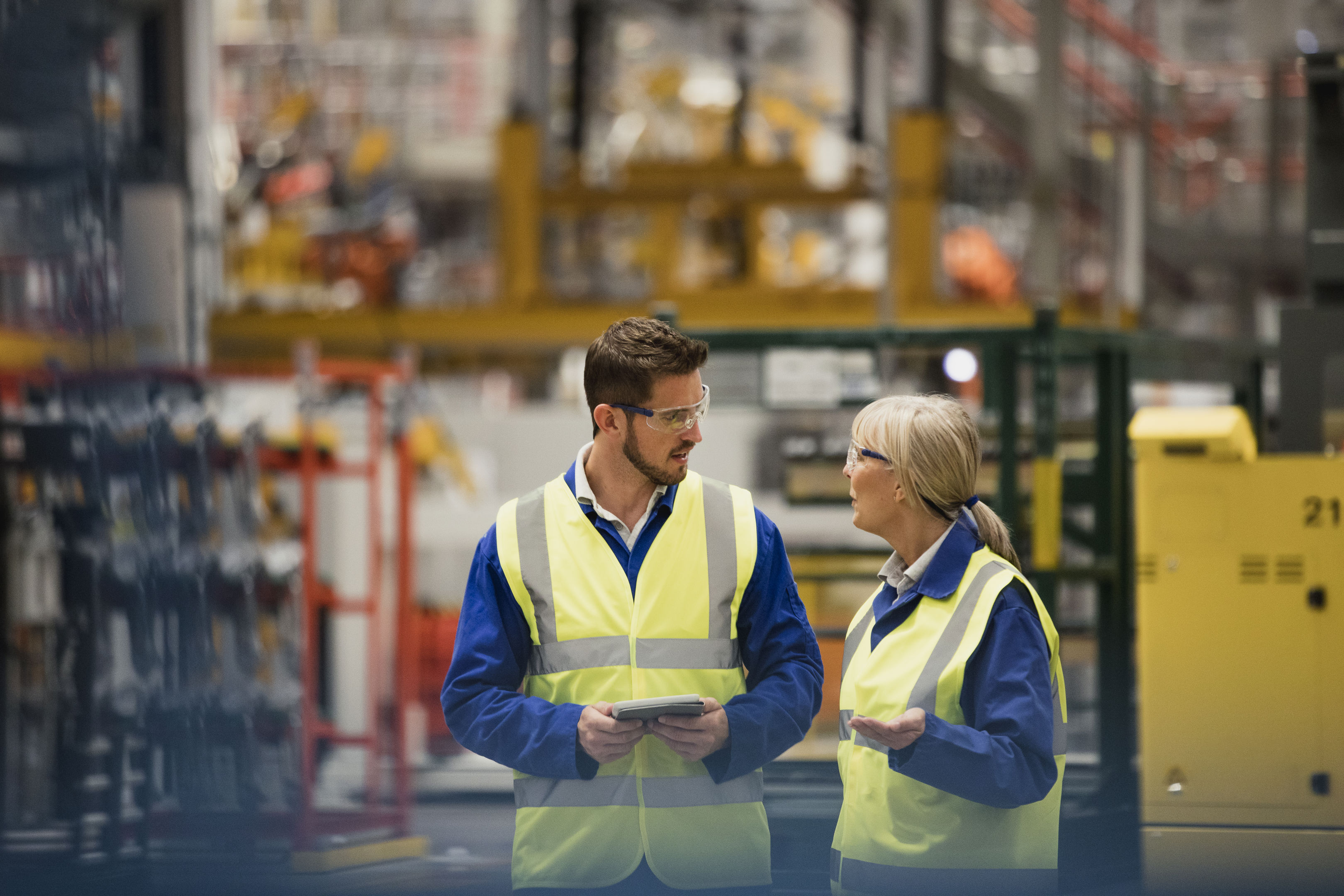 Two workers in high-visibility vests and safety glasses are talking in a large industrial facility. One holds a tablet while listening as the other gestures, with machinery and equipment visible in the background.