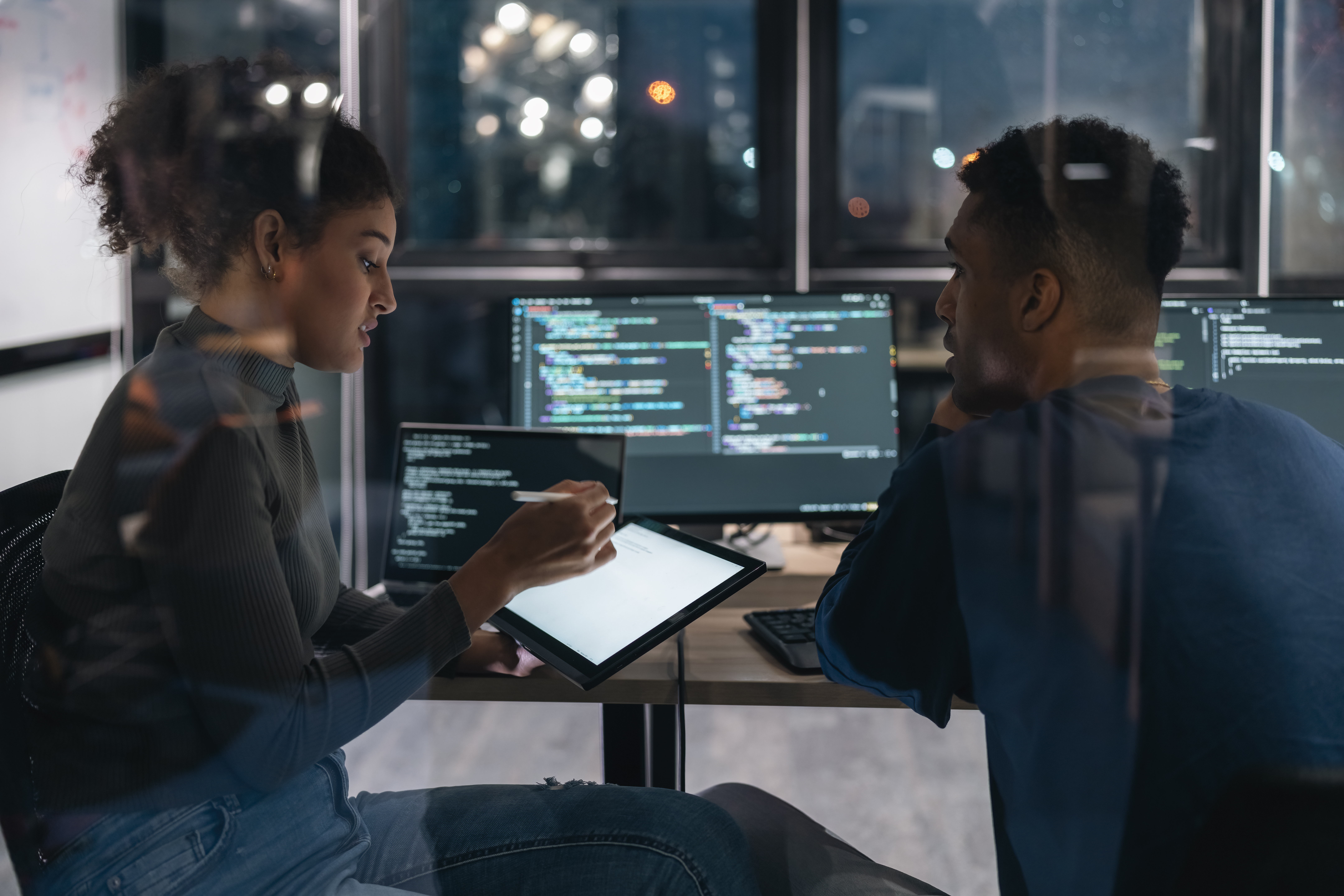 Two people sit at a desk with computer screens displaying code. One person gestures at a tablet, explaining something to the other, who listens attentively. The scene is in a modern office at night.