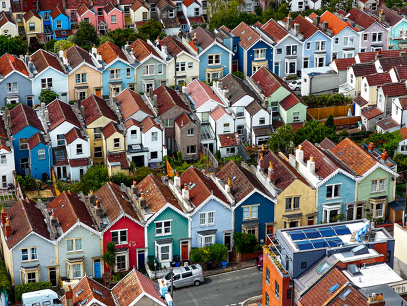 Aerial view of a neighborhood with rows of colorful houses, each with pitched roofs and small front gardens, lined along narrow streets with a few parked cars.