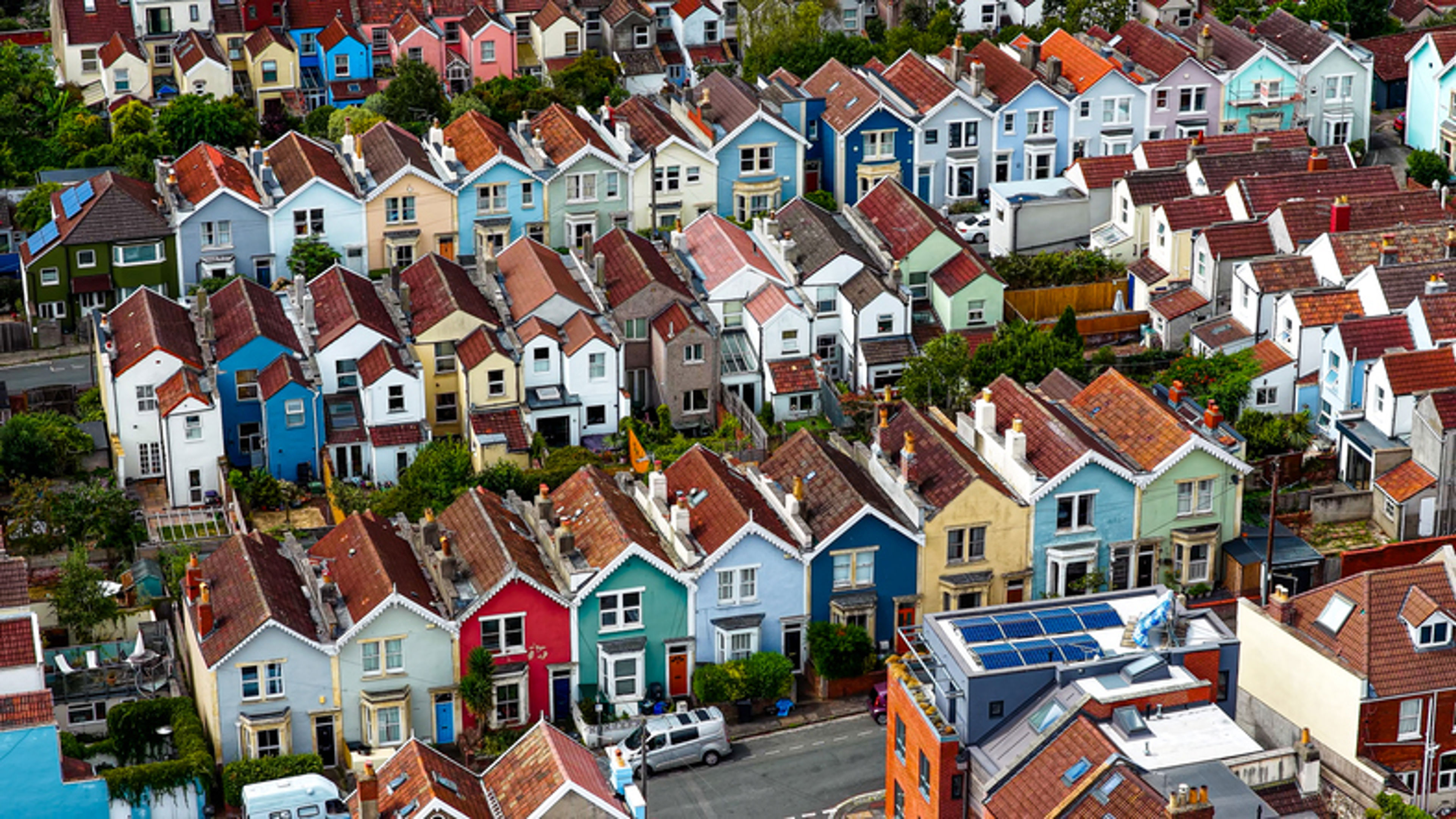 Aerial view of a neighborhood with rows of colorful houses, each with pitched roofs and small front gardens, lined along narrow streets with a few parked cars.