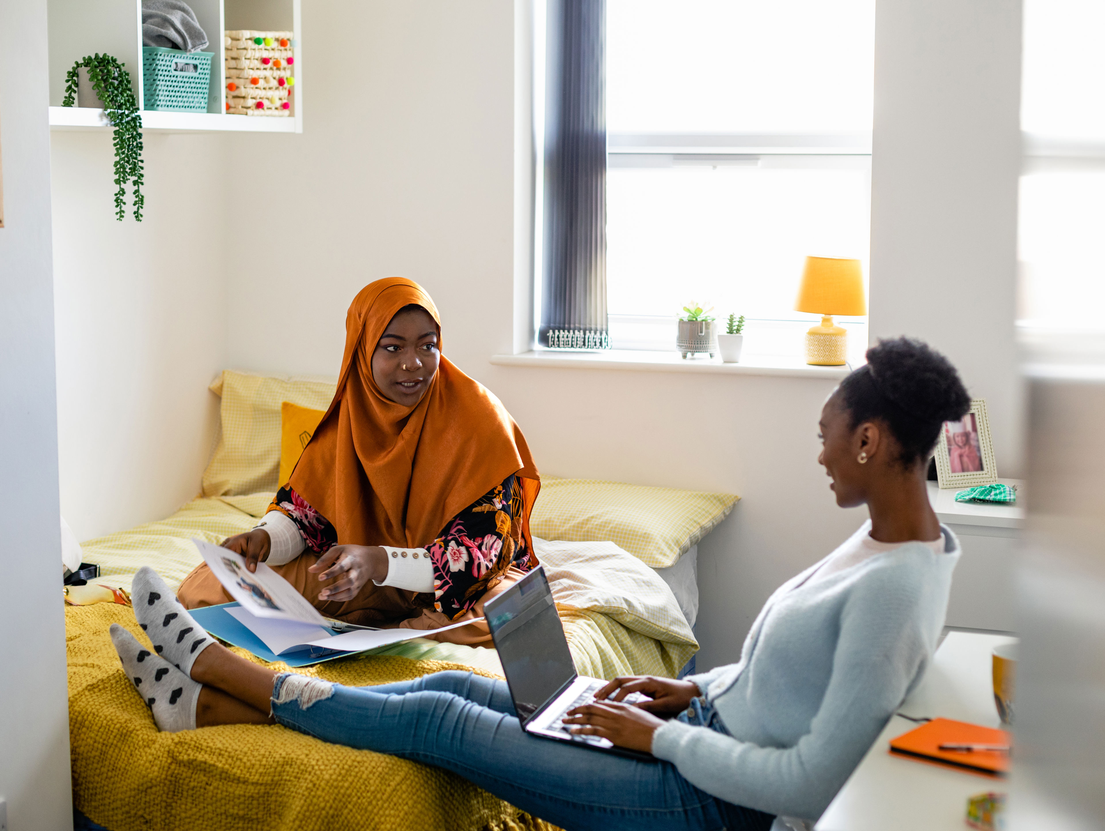 Two female uni student friends sitting in their dorm bedroom are studying together while sitting on the bed. They are using laptops/have their work files in front of them.