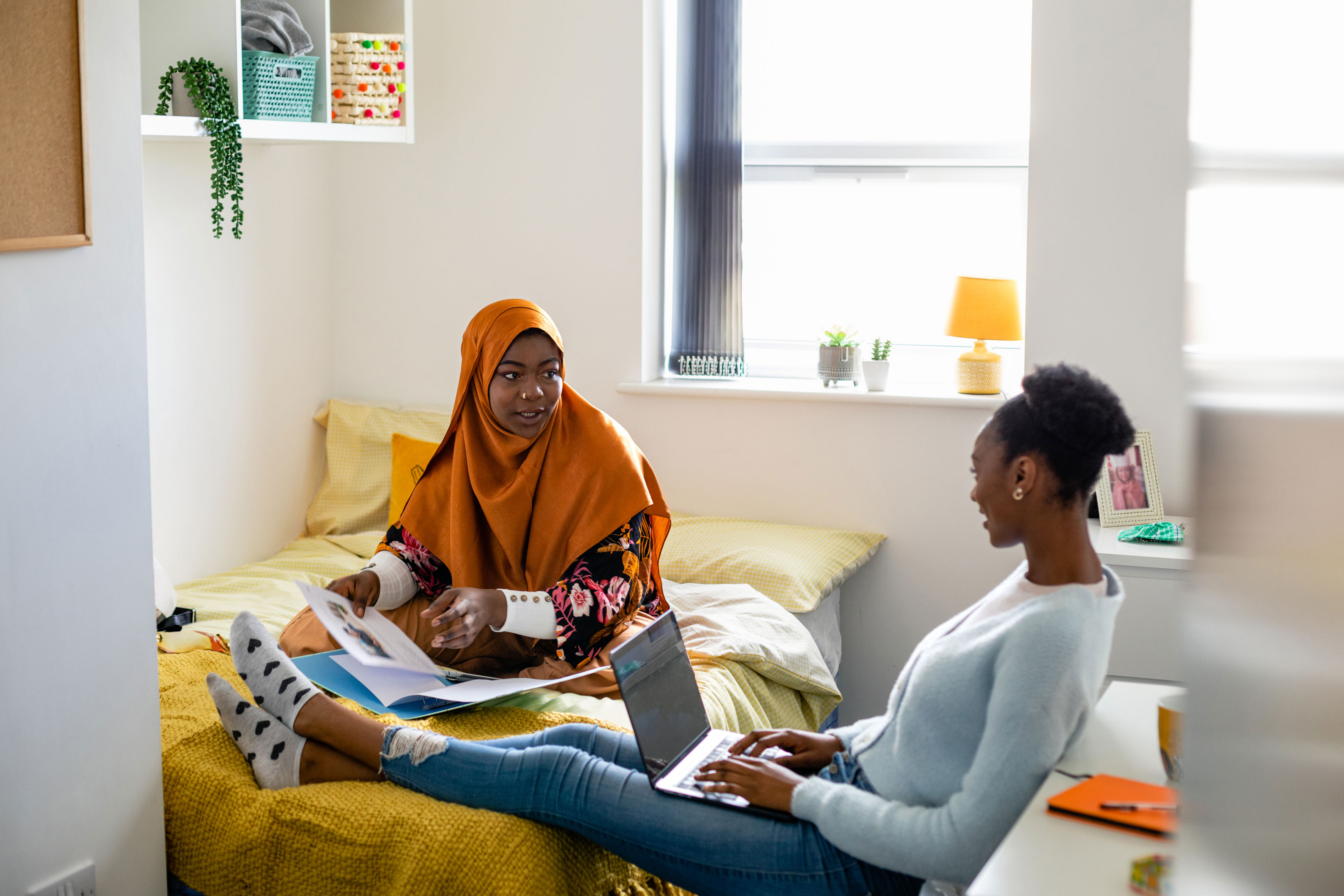 Two female uni student friends sitting in their dorm bedroom are studying together while sitting on the bed. They are using laptops/have their work files in front of them.