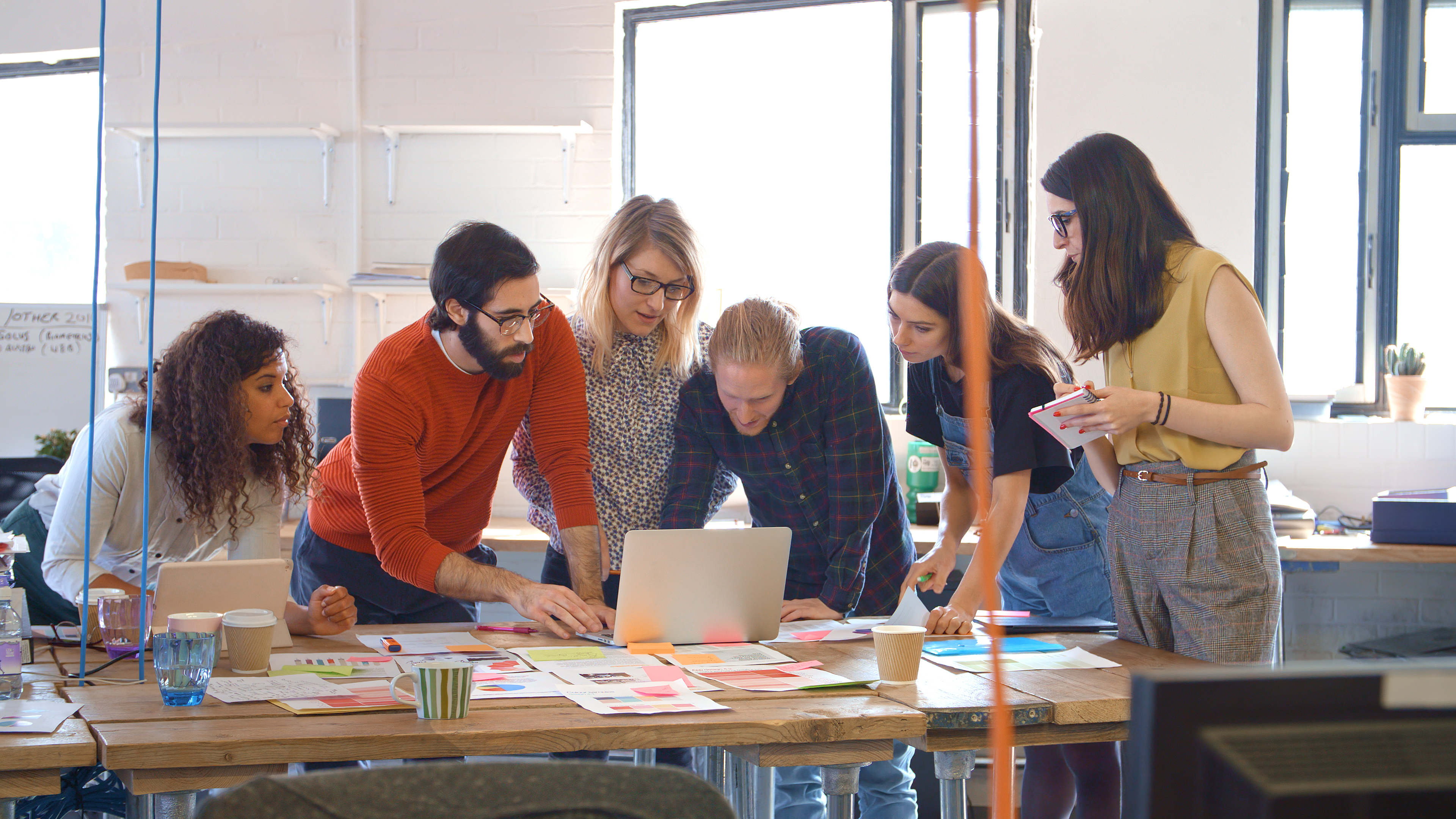 Six people gather around a table, looking at a laptop and discussing documents and charts spread out, in a bright modern office setting.