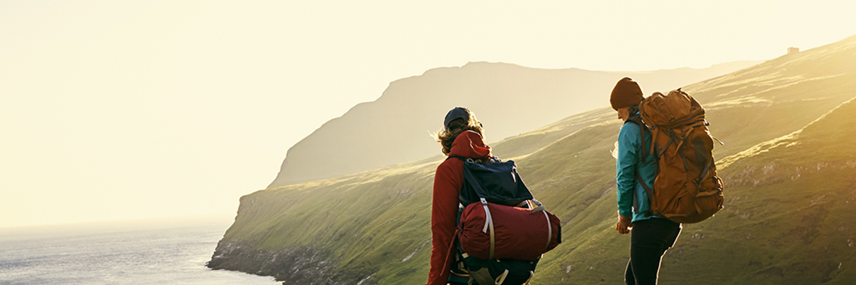 Two hikers with backpacks walk along a grassy hill overlooking a coastal cliff and the ocean at sunset, with sunlight illuminating the landscape and distant mountains in the background.