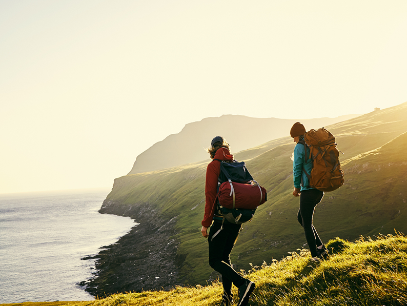 Two hikers with backpacks walk along a grassy hill overlooking a coastal cliff and the ocean at sunset, with sunlight illuminating the landscape and distant mountains in the background.