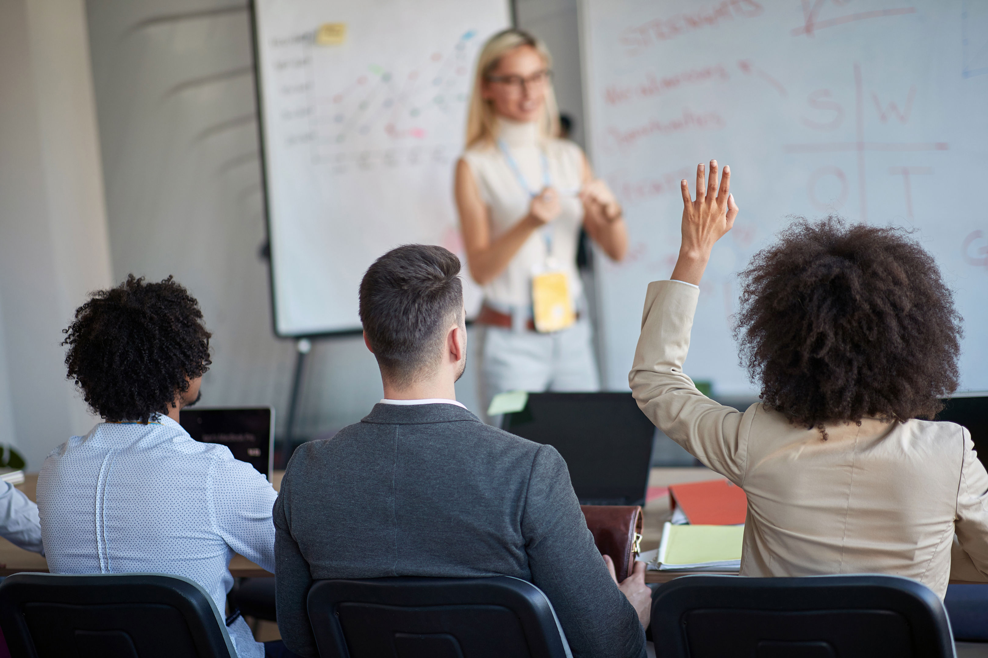 A group of people sit facing a woman presenting in front of a whiteboard. One attendee raises their hand, while others listen and take notes. The whiteboard has diagrams and text.
