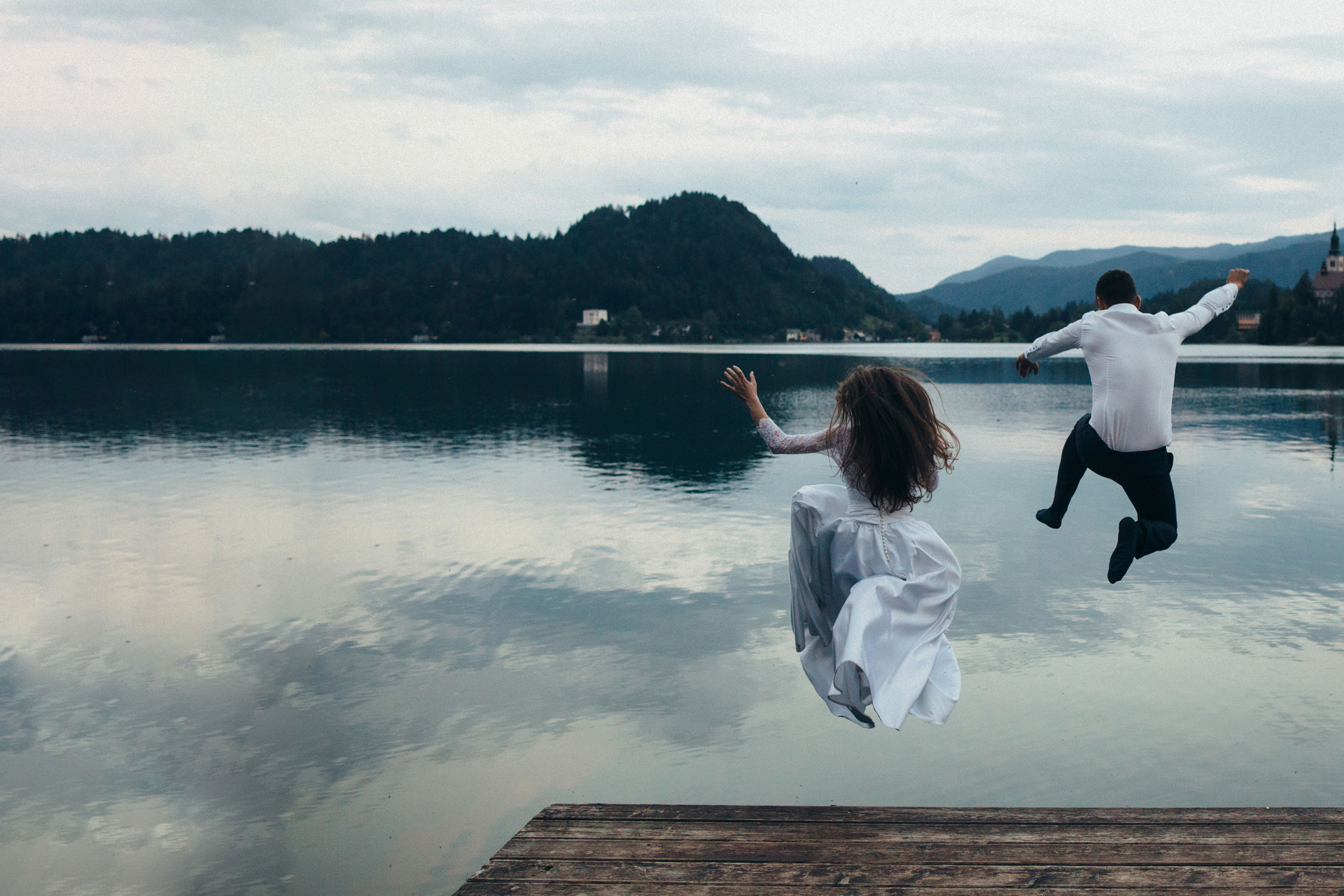 A bride and groom, dressed in wedding attire, joyfully jump off a wooden dock into a calm lake, with mountains and cloudy skies in the background.