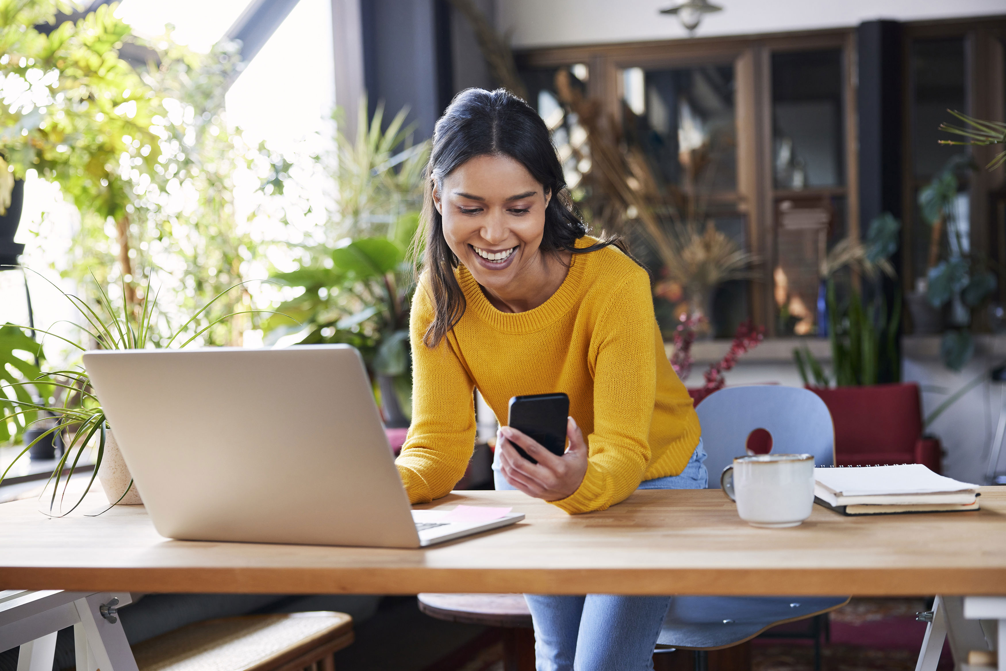Smiling person in a yellow sweater looks at a smartphone while leaning on a desk with a laptop, notebook, and mug. The room has large windows and is decorated with plants.