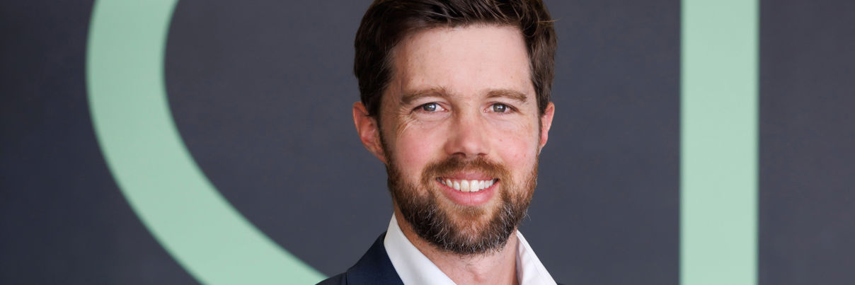 A smiling man with a beard wearing a dark blue suit and white button-down shirt stands in front of a dark wall with a large green graphic.