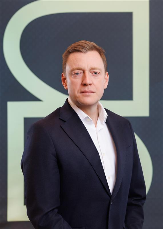 A man in a dark suit and white dress shirt stands in front of a geometric background featuring large, light-colored shapes. He is looking directly at the camera with a neutral expression.