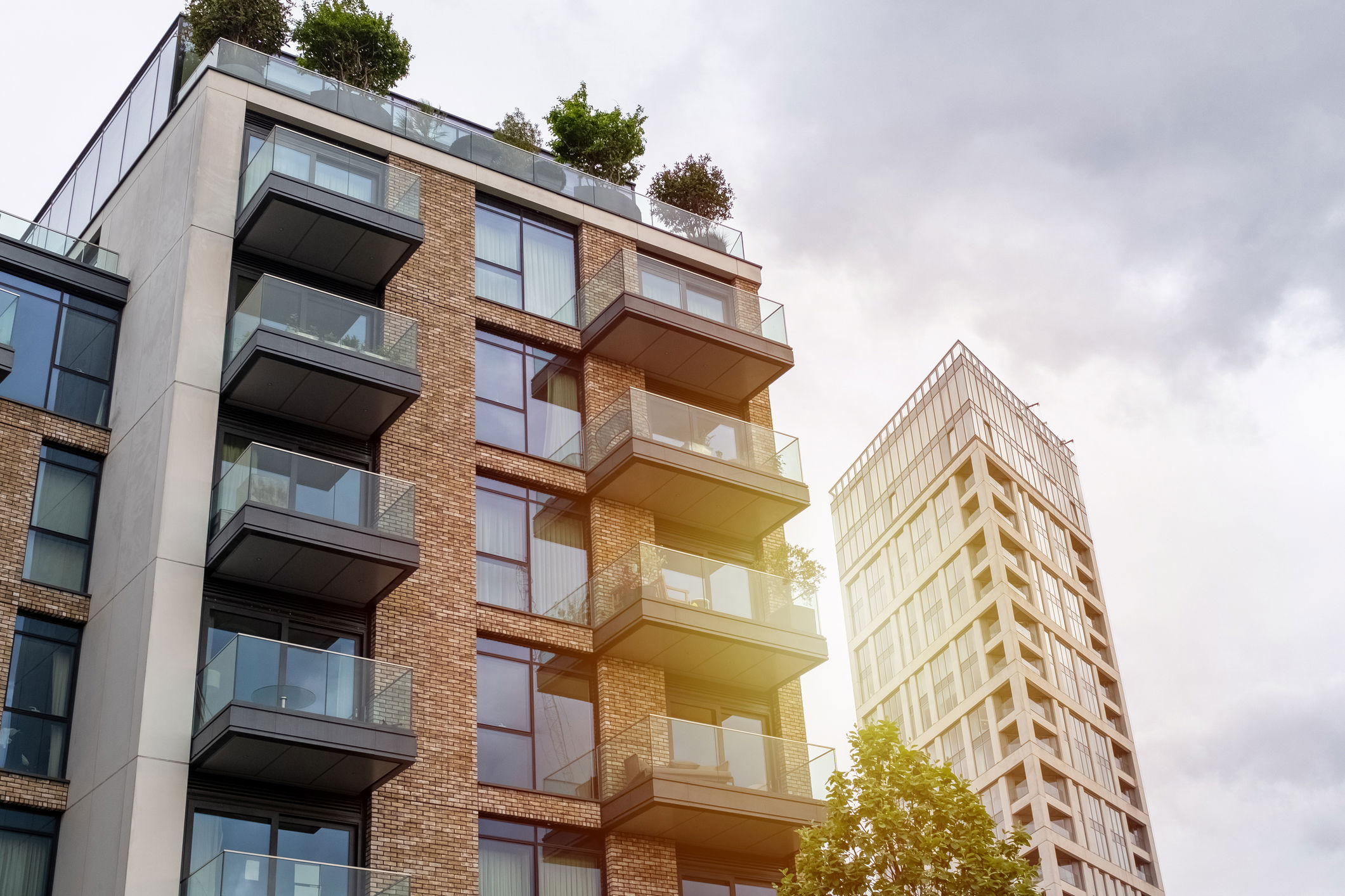 A modern apartment building with brick and glass balconies is in the foreground. A taller building with a geometric facade is visible in the background. The sky is overcast with a burst of sunlight peeking through. Trees are on some balconies.