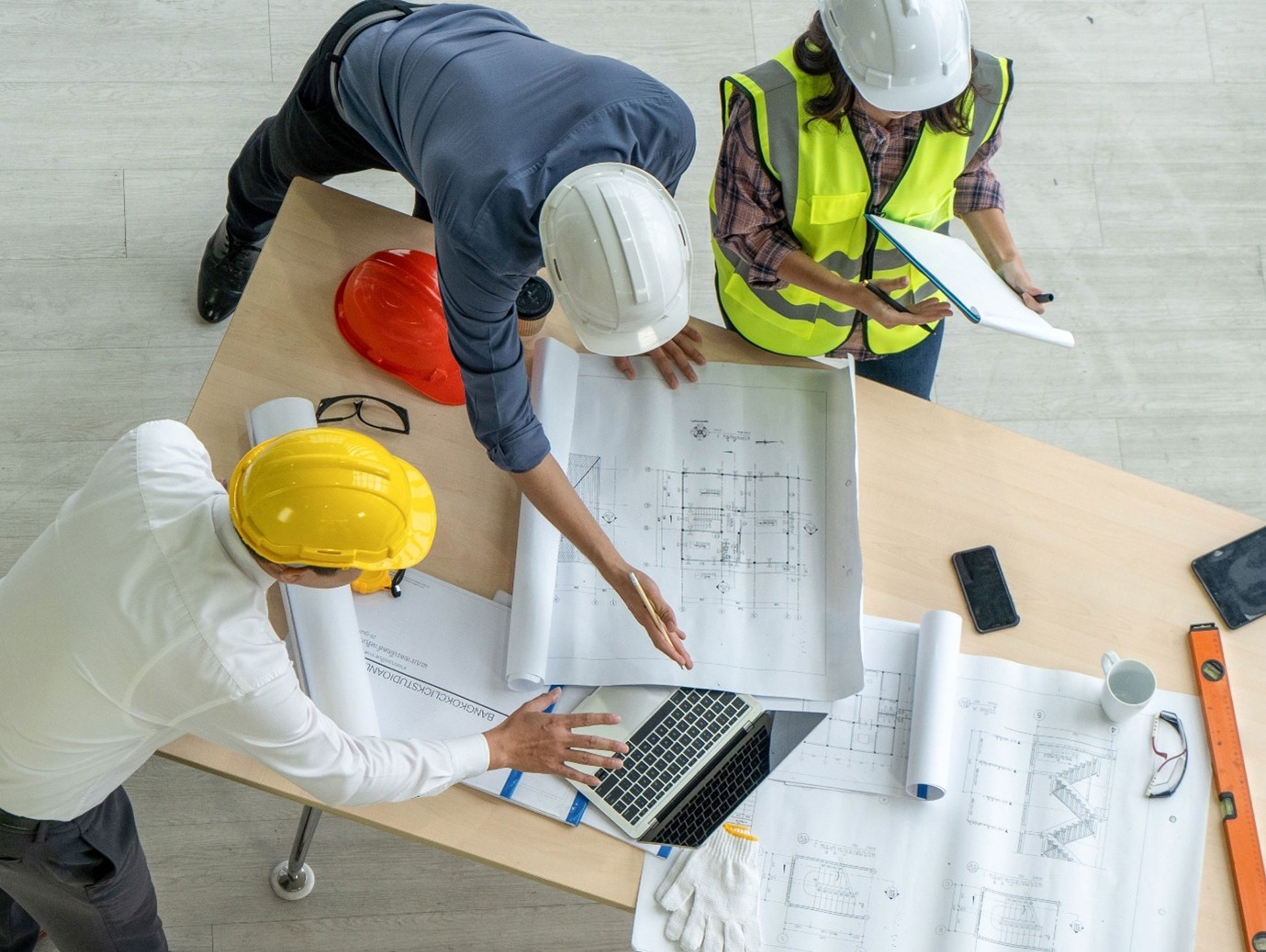 Three construction professionals wearing safety helmets and vests review blueprints and documents on a table with a laptop, hard hats, gloves, and tools visible. Photographed from above.