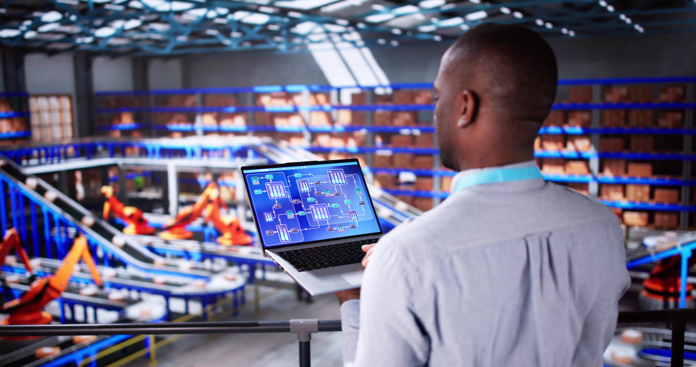 A man stands overlooking an automated warehouse while holding a laptop displaying a digital schematic or system diagram. Conveyor belts, shelves, and robotic arms are visible in the large, modern facility.