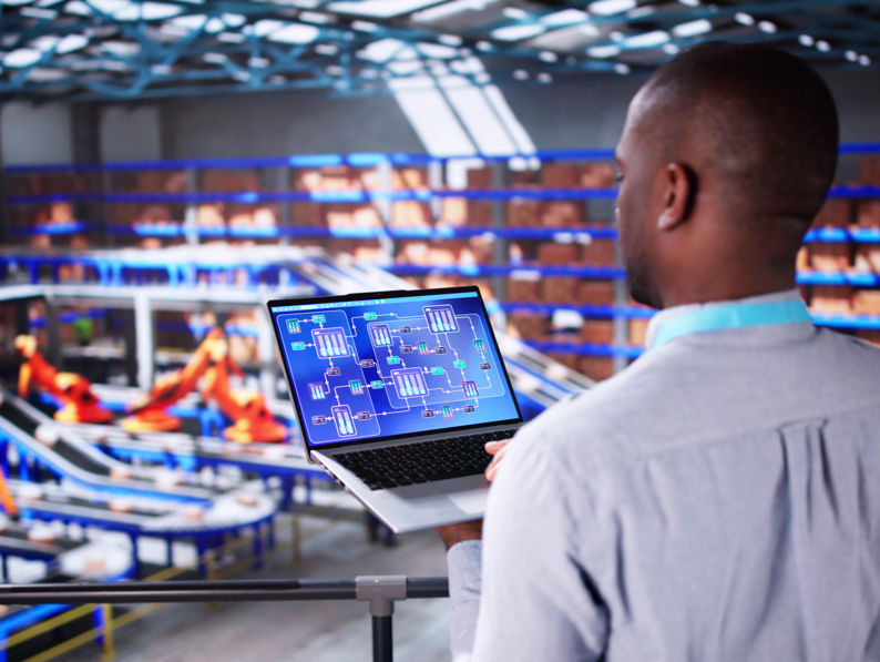 A man stands overlooking an automated warehouse while holding a laptop displaying a digital schematic or system diagram. Conveyor belts, shelves, and robotic arms are visible in the large, modern facility.