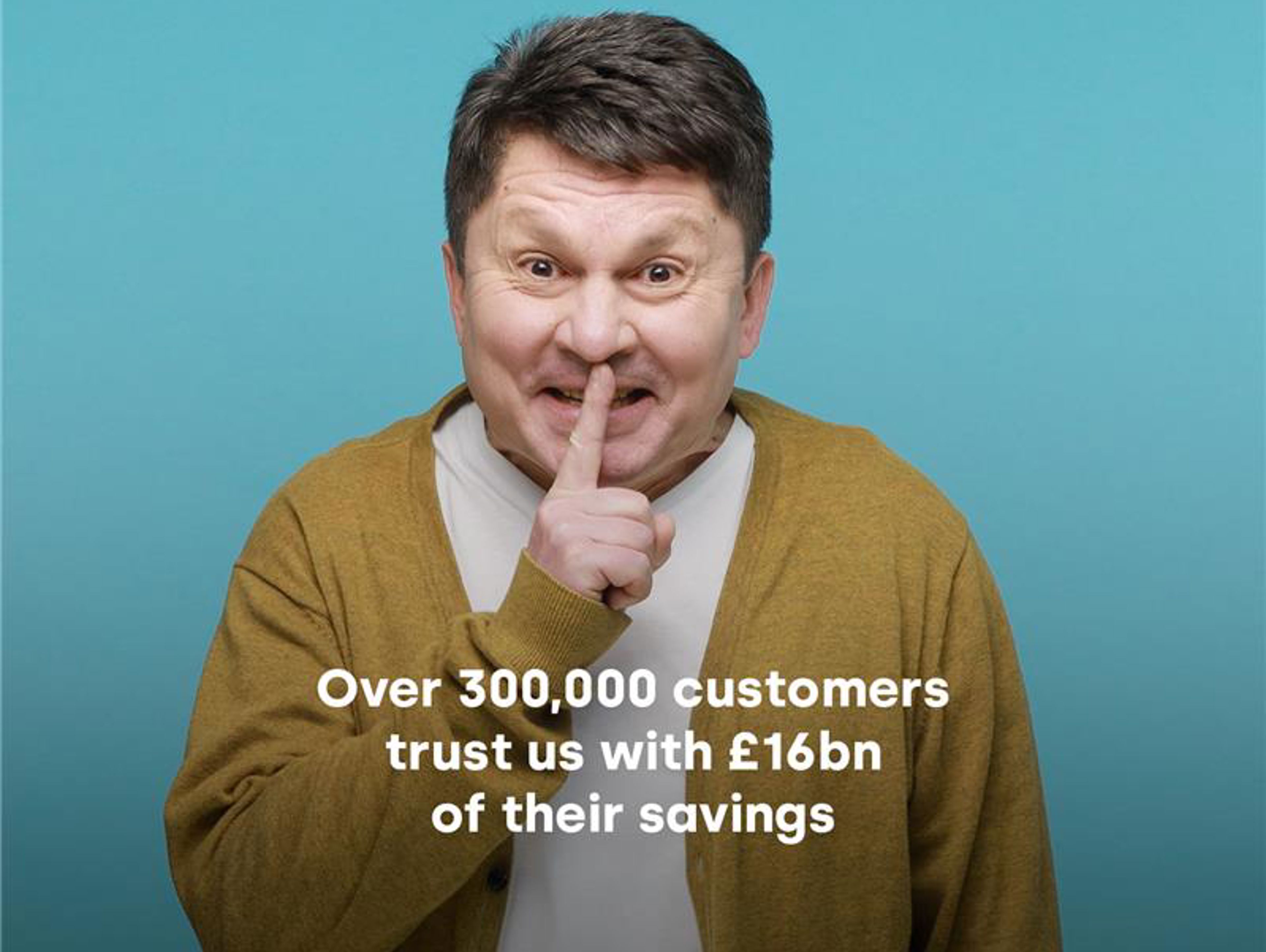 A man in a mustard cardigan holds a finger to his lips, signaling "shh." Text reads: "Over 300,000 customers trust us with £16bn of their savings." The background is plain blue.