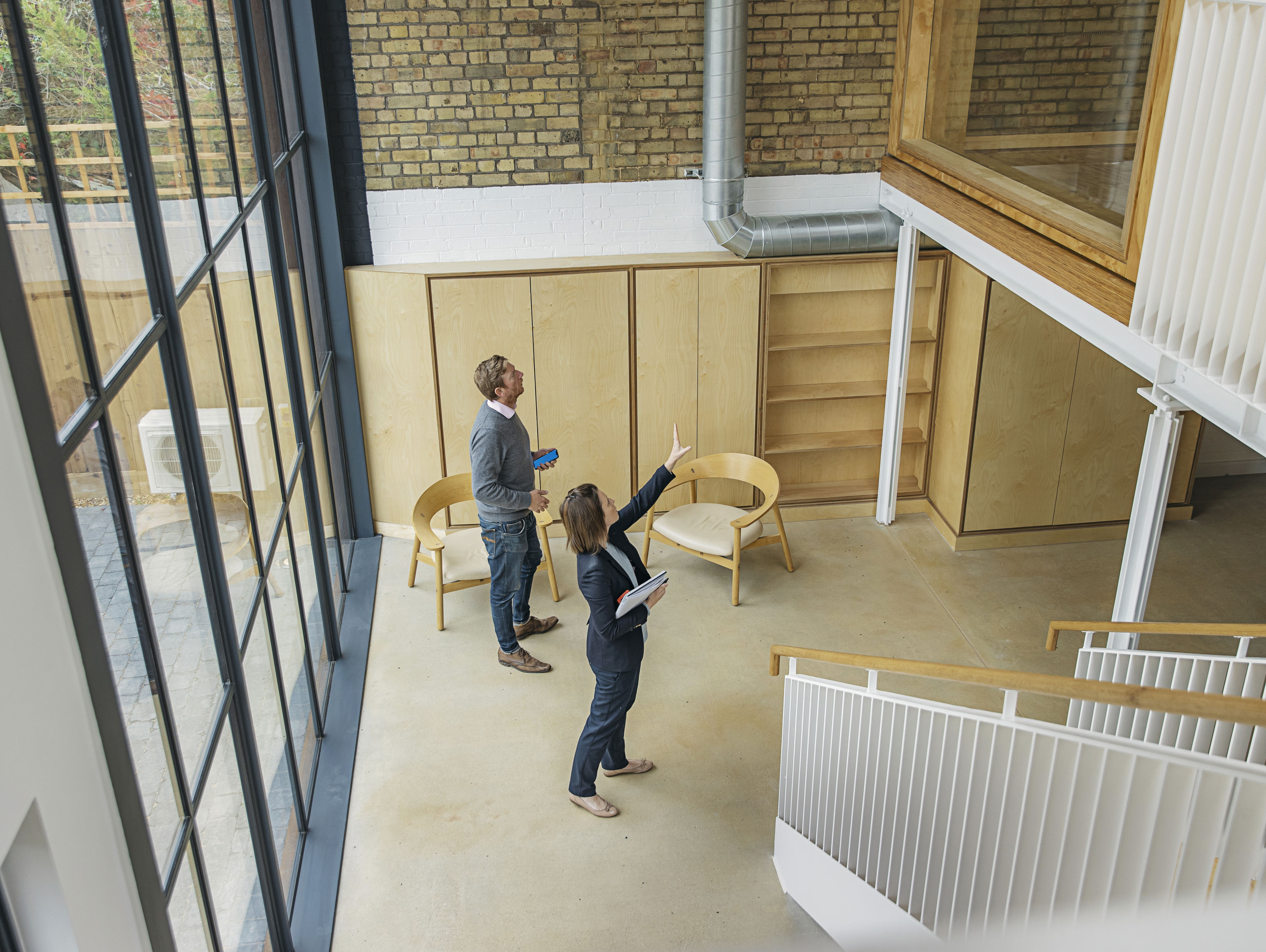 Two people stand in a modern, bright room with large windows, wooden walls, and minimalist furniture. One person points upwards, possibly discussing the space, while the other listens and holds a tablet.