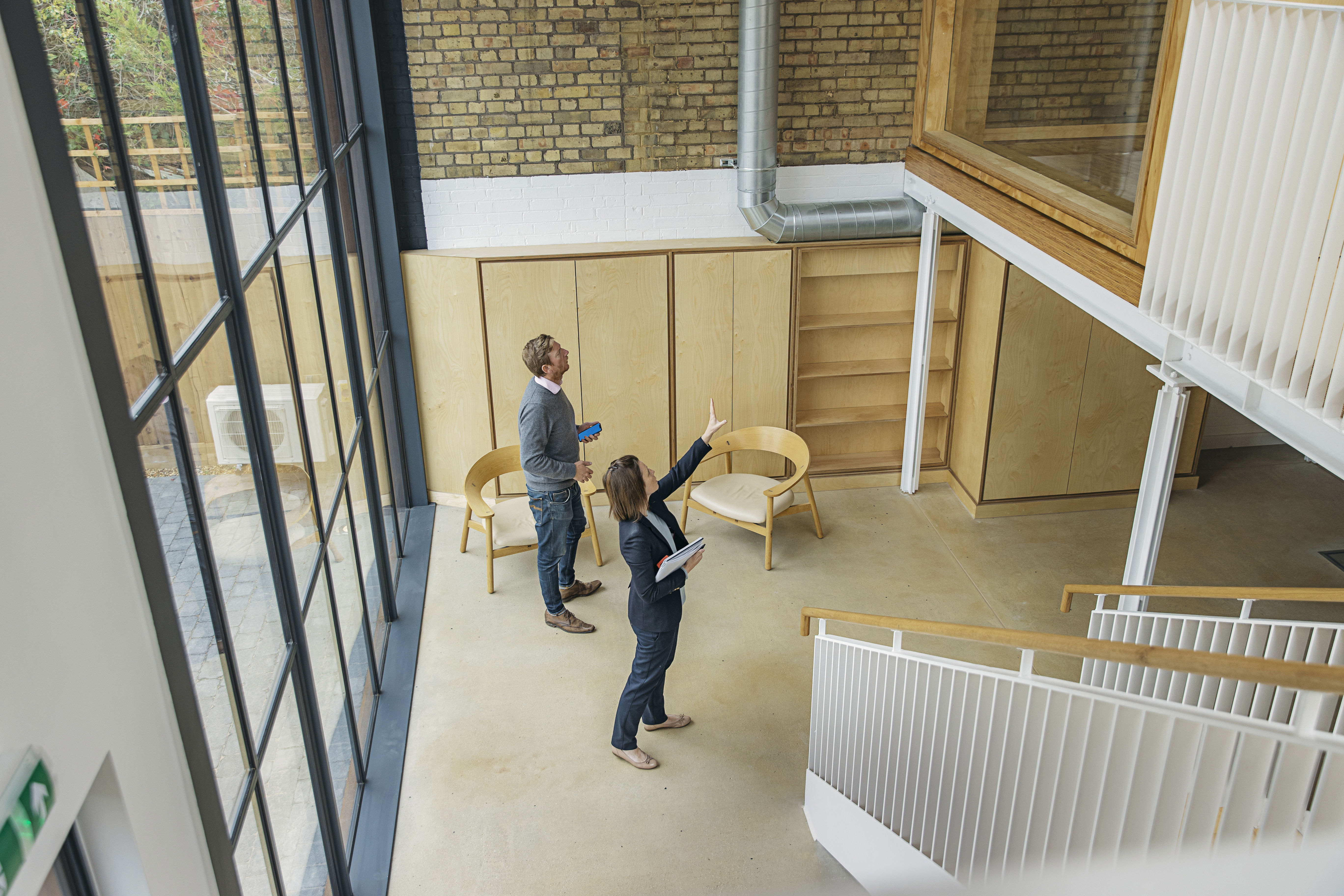 Two people stand in a modern, bright room with large windows, wooden walls, and minimalist furniture. One person points upwards, possibly discussing the space, while the other listens and holds a tablet.
