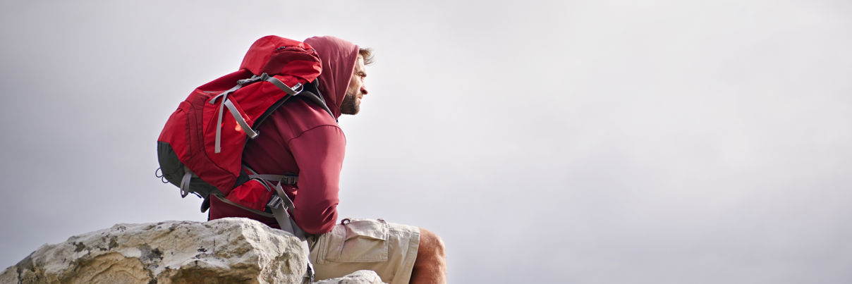 A person wearing a red hoodie and backpack sits on a rocky outcrop, looking thoughtfully into the distance under a cloudy sky.