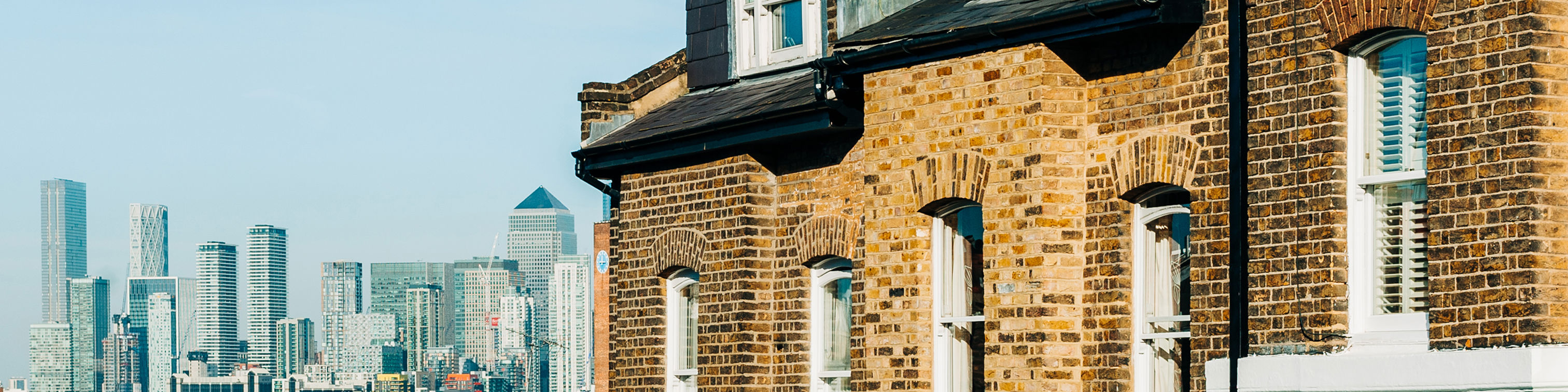 Row of traditional brick houses with pastel-colored facades on the right, set against a clear blue sky. In the background, modern skyscrapers of a city skyline are visible, creating a contrast between old and new architecture.