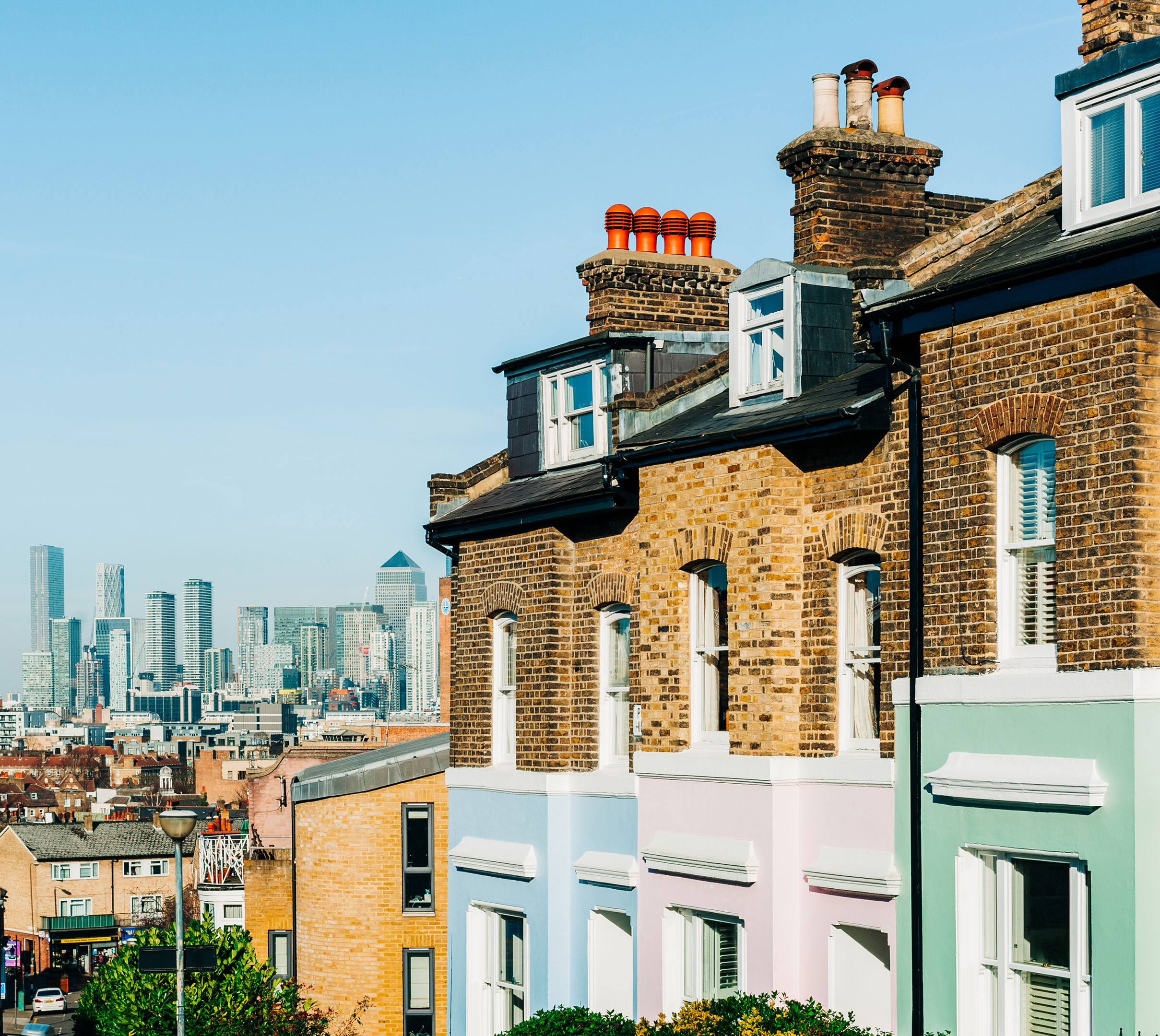 Row of traditional brick houses with pastel-colored facades on the right, set against a clear blue sky. In the background, modern skyscrapers of a city skyline are visible, creating a contrast between old and new architecture.