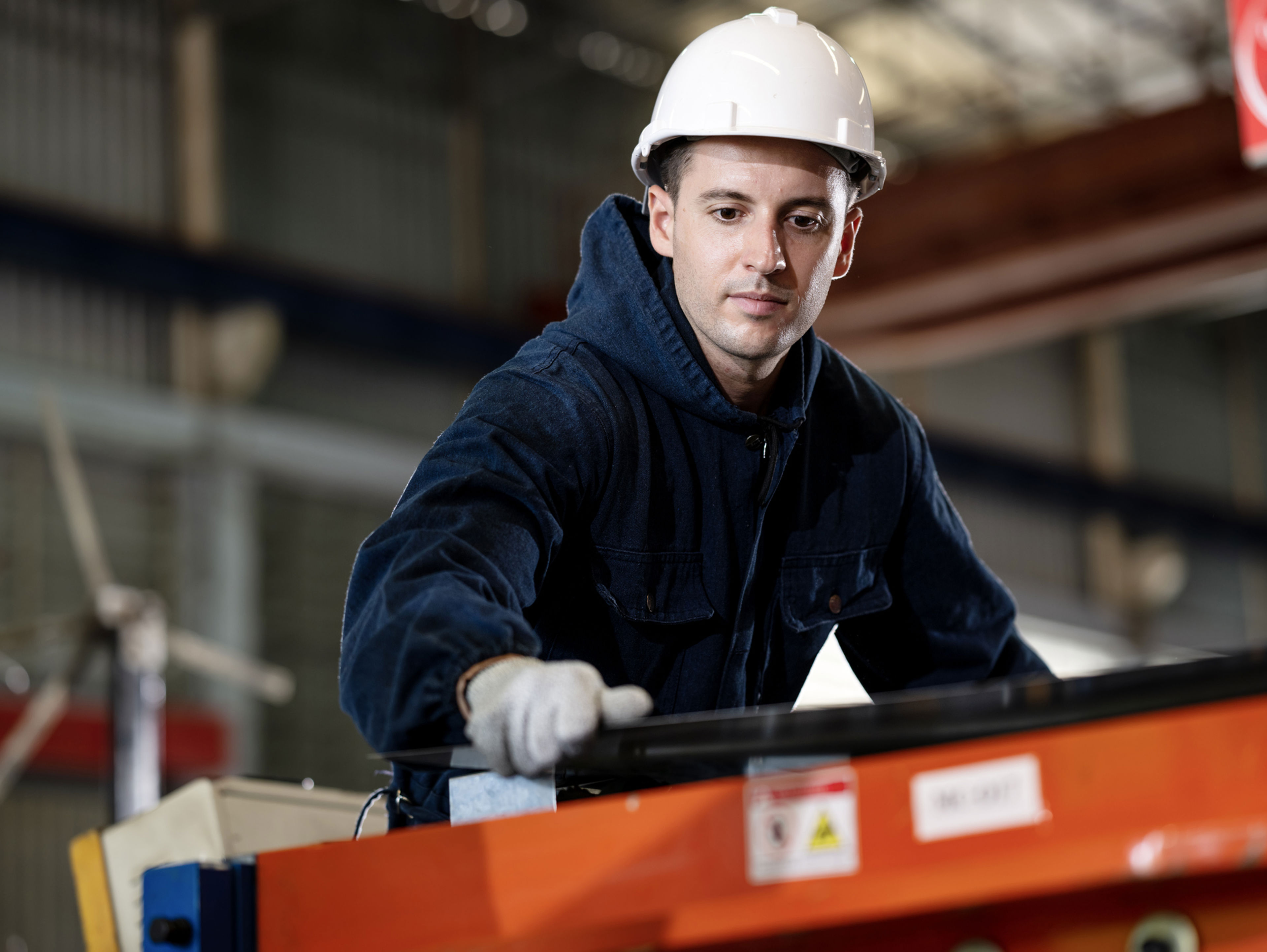 A worker in a dark blue uniform and white hard hat operates machinery in an industrial setting, focusing intently while using a gloved hand to guide materials.
