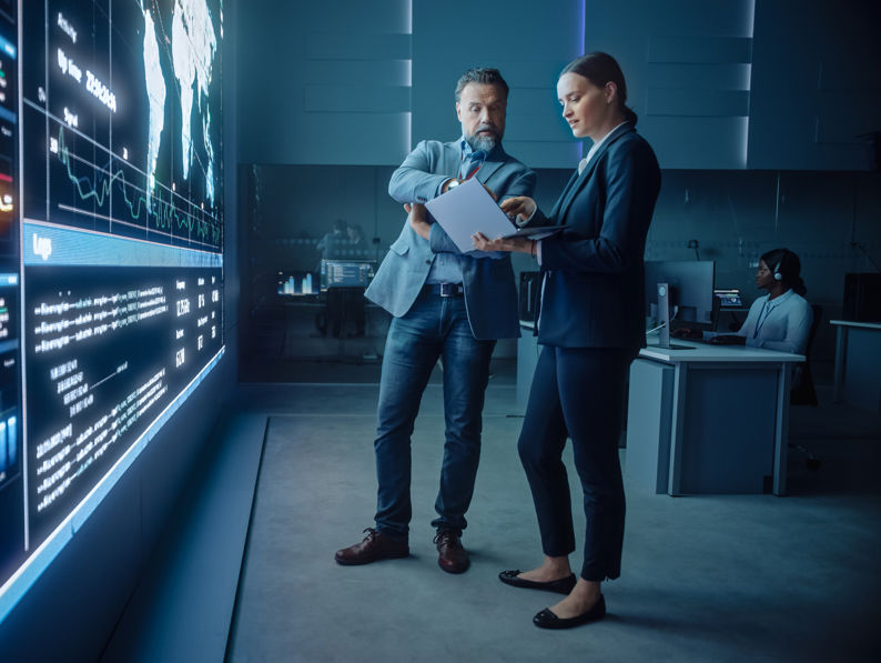 Project Manager and Computer Science Engineer Talk while Using Big Screen Display and a Laptop, Showing Infrastructure Infographics Data. Telecommunications Company System Control and Monitoring Room