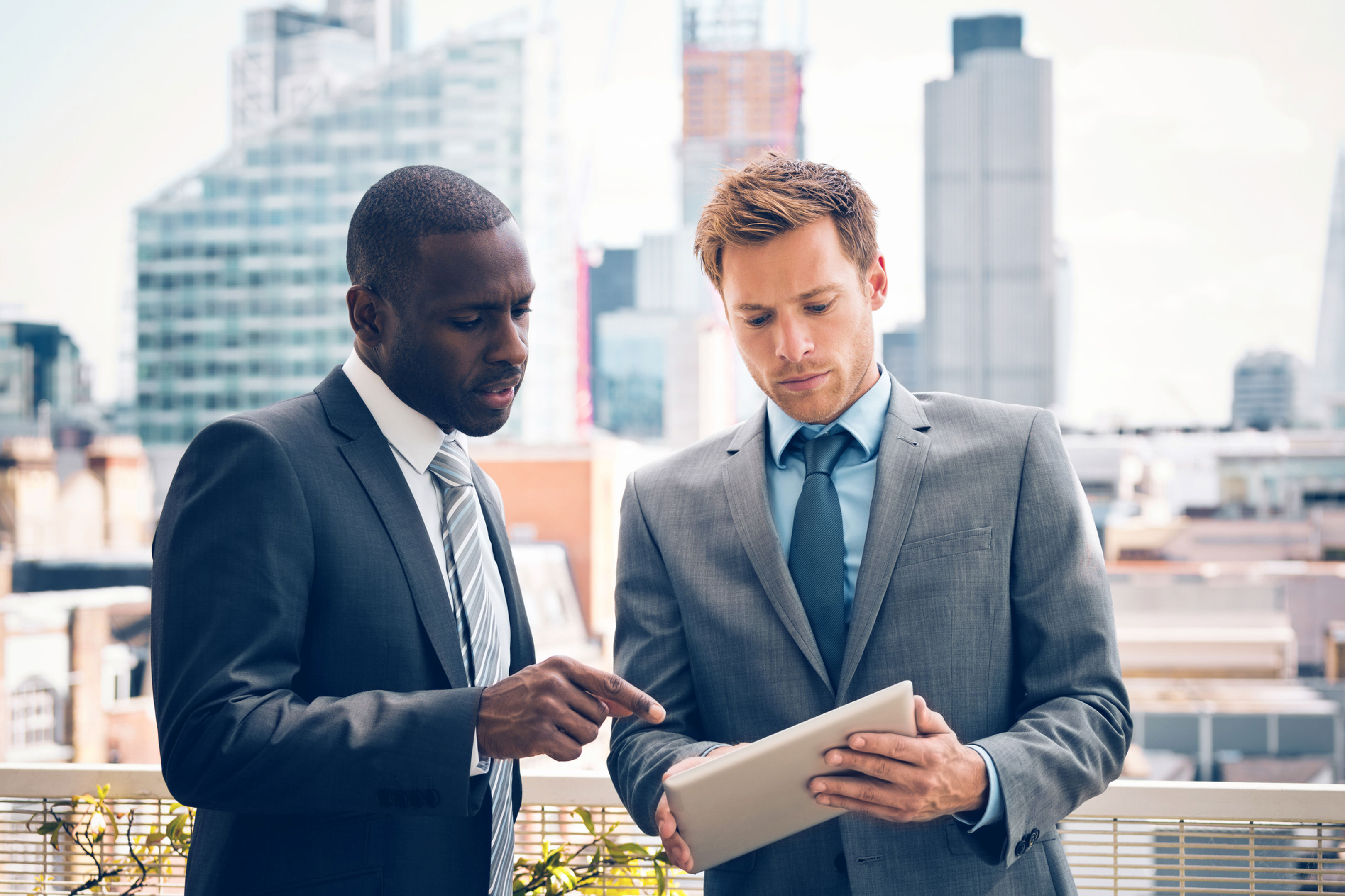 Two businessmen in suits stand on a rooftop with a city skyline in the background. One man is holding a tablet while the other points at the screen, both focused on the device, engaged in discussion.