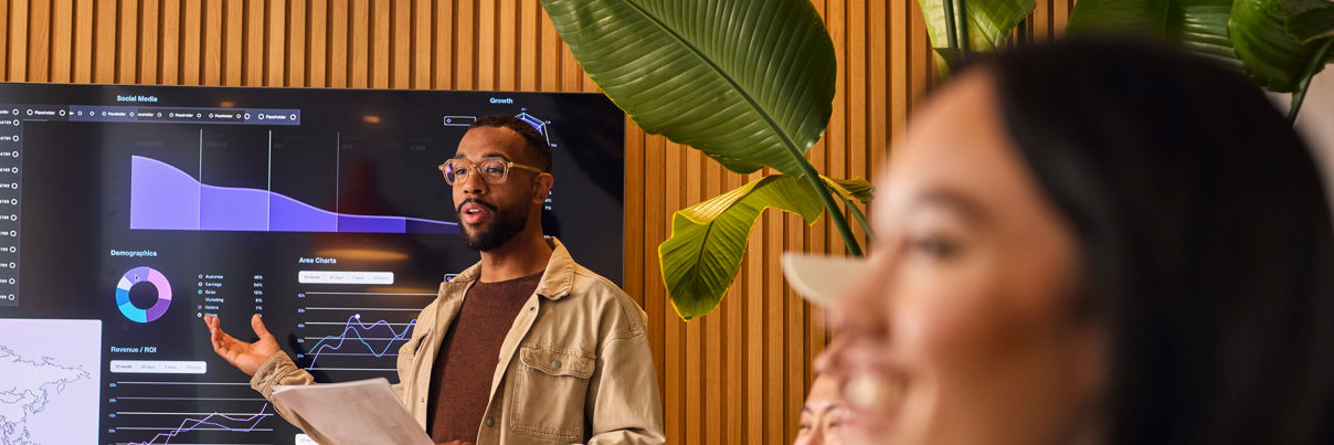 A man stands in front of a digital screen displaying charts and graphs, presenting data to a group of colleagues seated at a table in a modern office with wooden walls and large green plants.