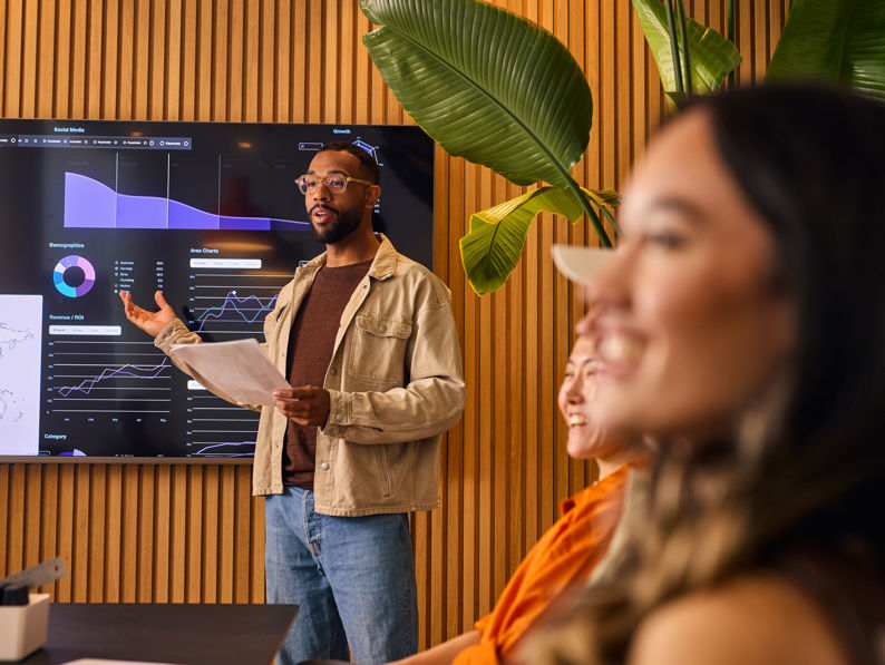 A man stands in front of a digital screen displaying charts and graphs, presenting data to a group of colleagues seated at a table in a modern office with wooden walls and large green plants.