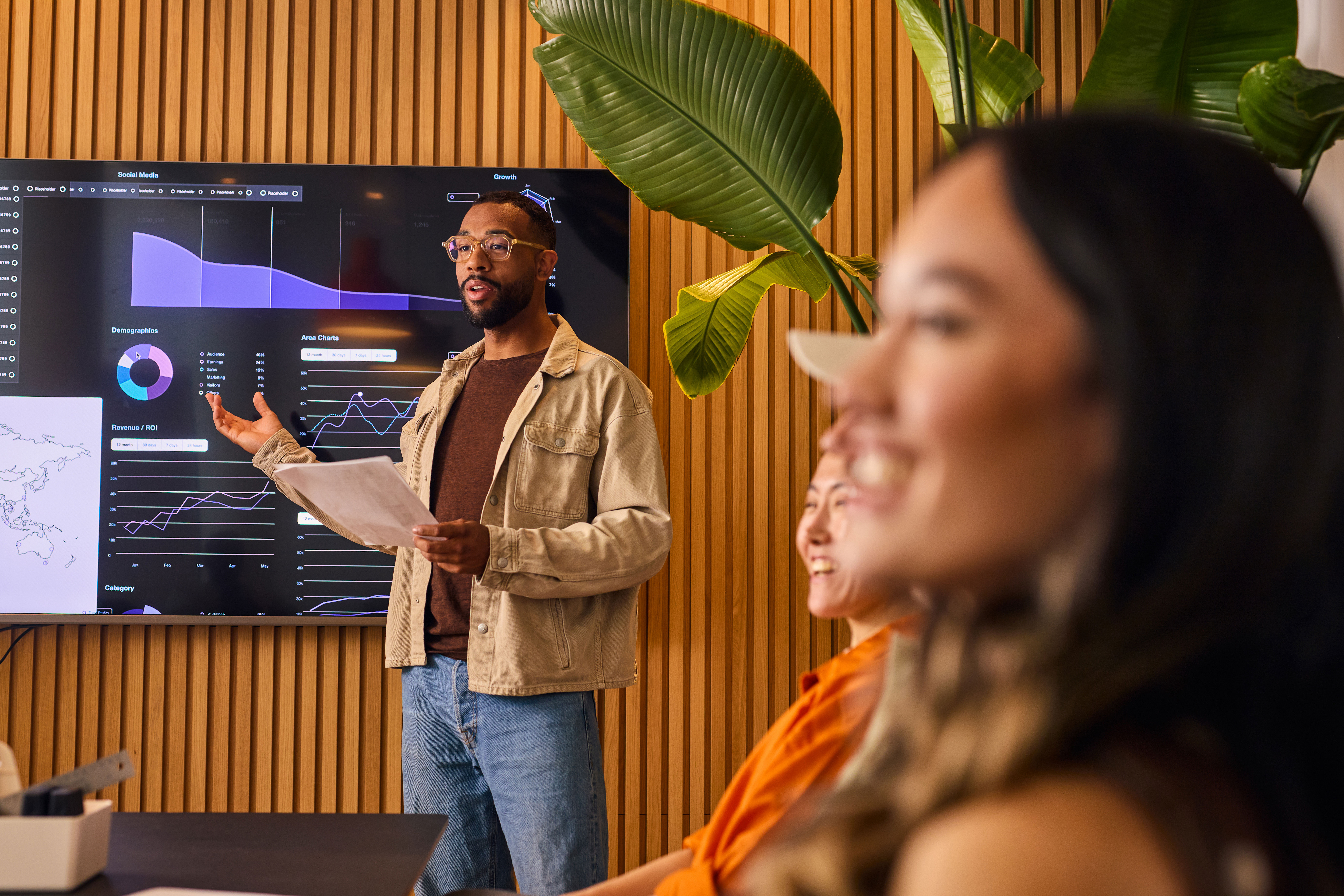 A man stands in front of a digital screen displaying charts and graphs, presenting data to a group of colleagues seated at a table in a modern office with wooden walls and large green plants.
