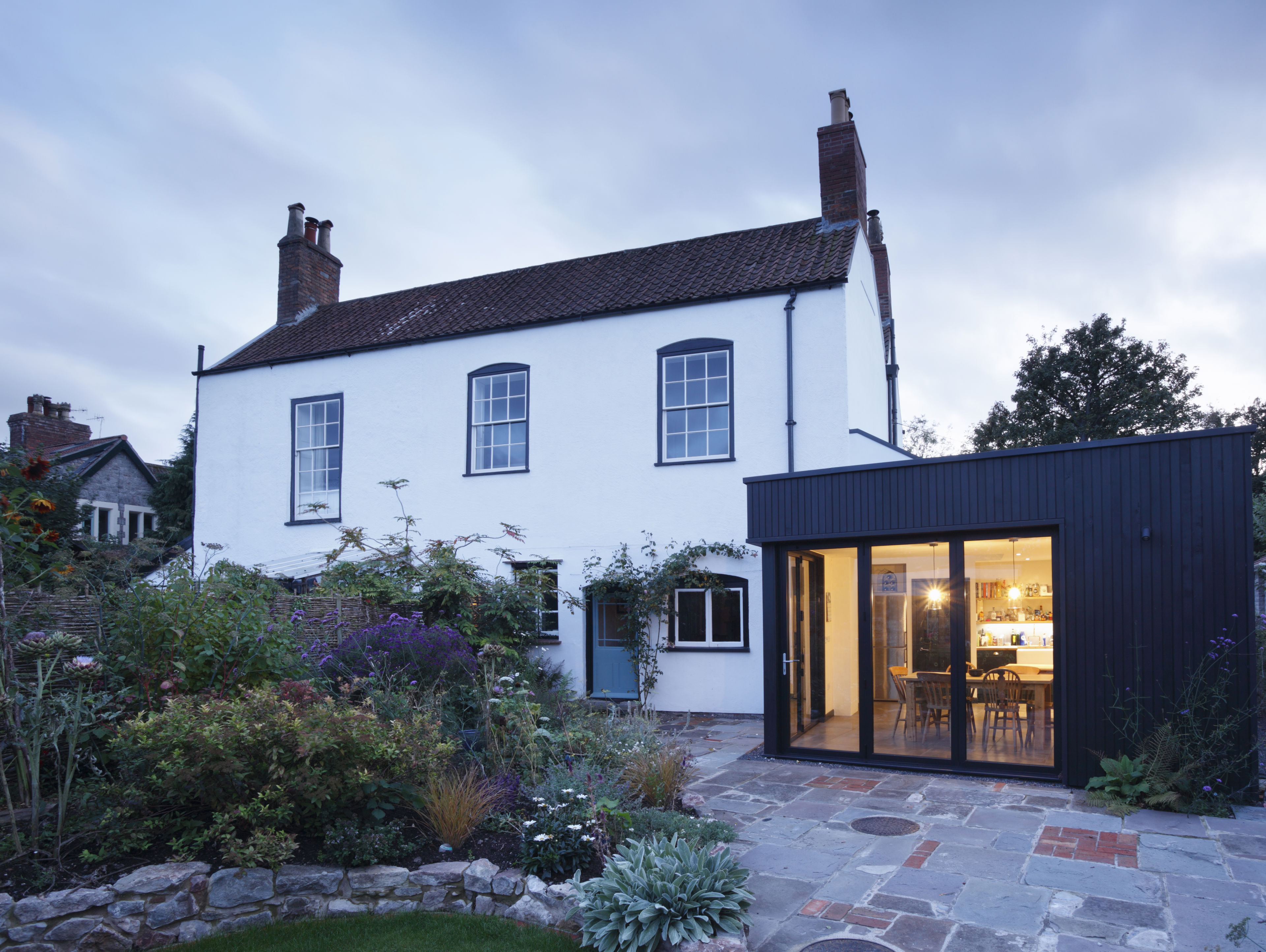 A traditional white two-story house with a red-tiled roof stands beside a modern black extension with large windows, revealing a well-lit dining area inside. A stone patio and garden are in the foreground.
