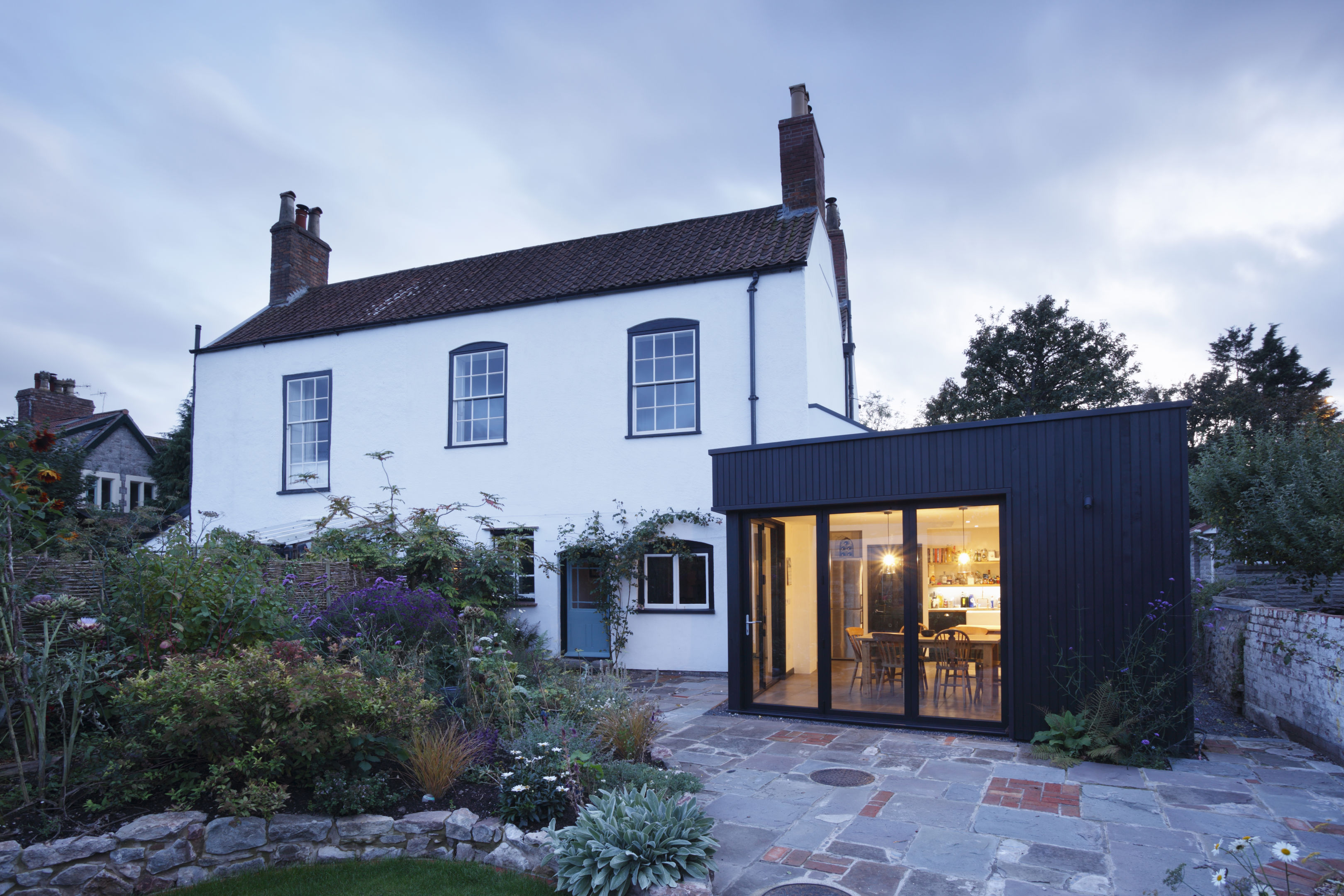 A traditional white two-story house with a red-tiled roof stands beside a modern black extension with large windows, revealing a well-lit dining area inside. A stone patio and garden are in the foreground.