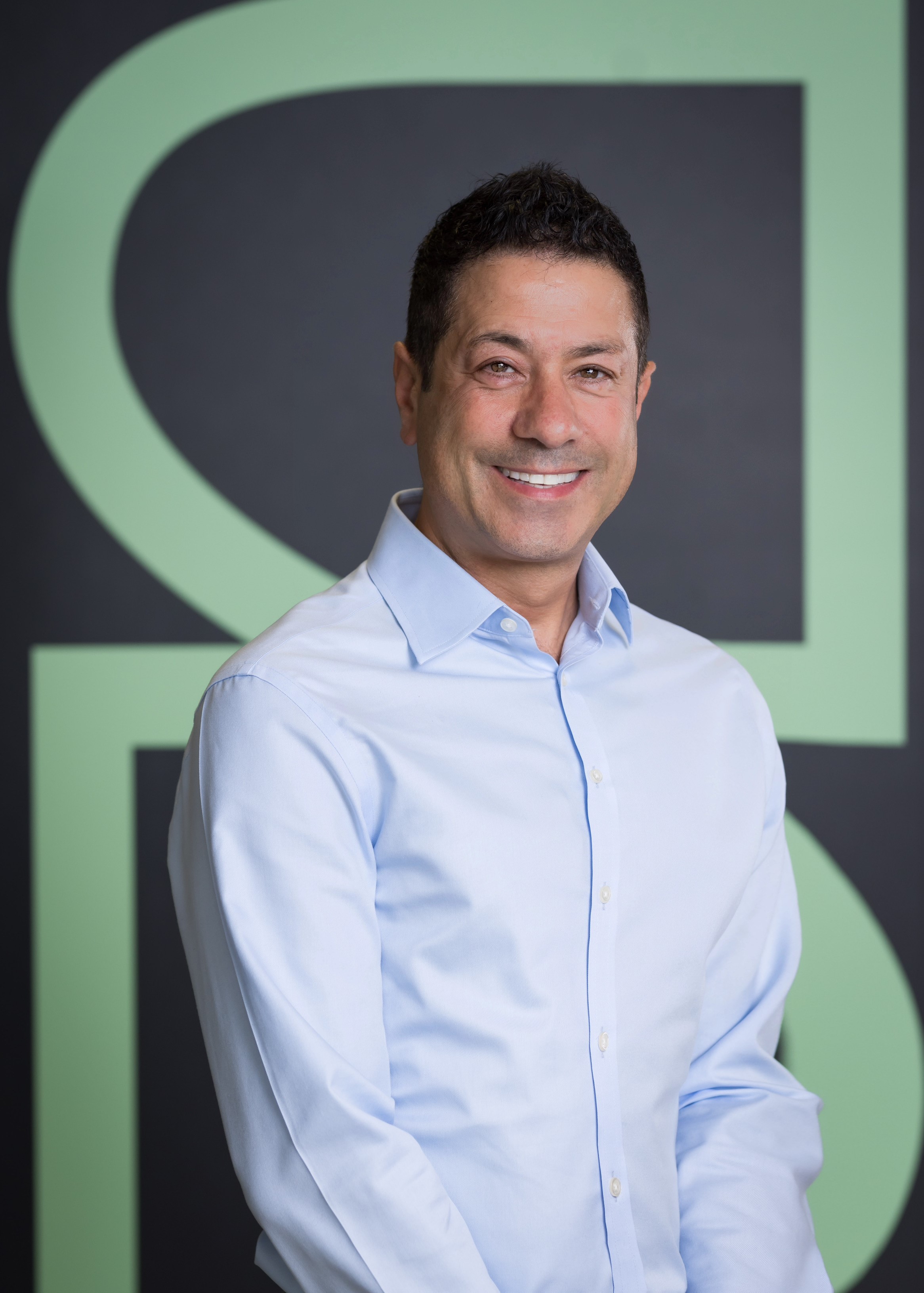 Smiling man in a light blue shirt stands against a dark background with large, light green letters.