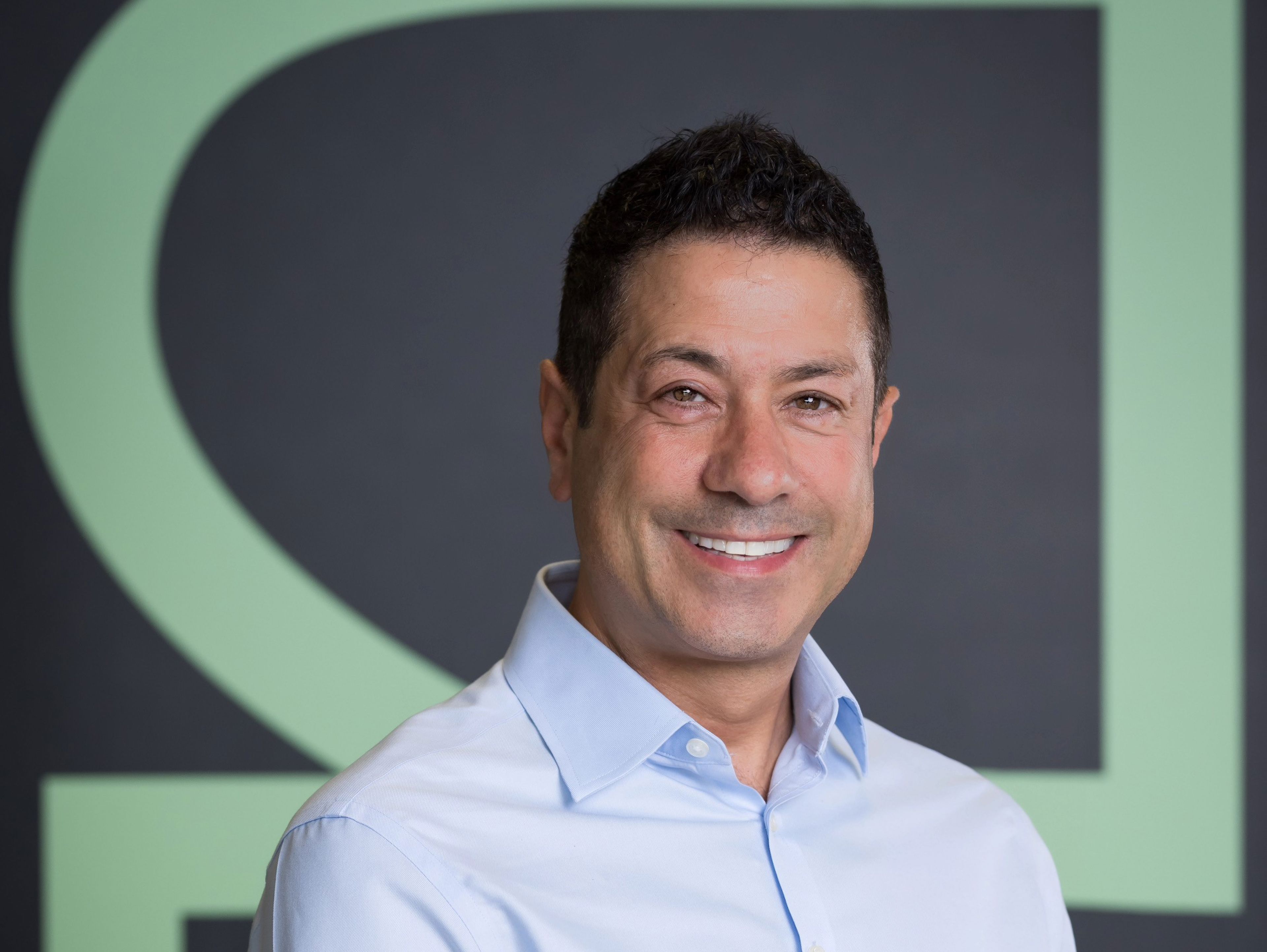 Smiling man in a light blue shirt stands against a dark background with large, light green letters.