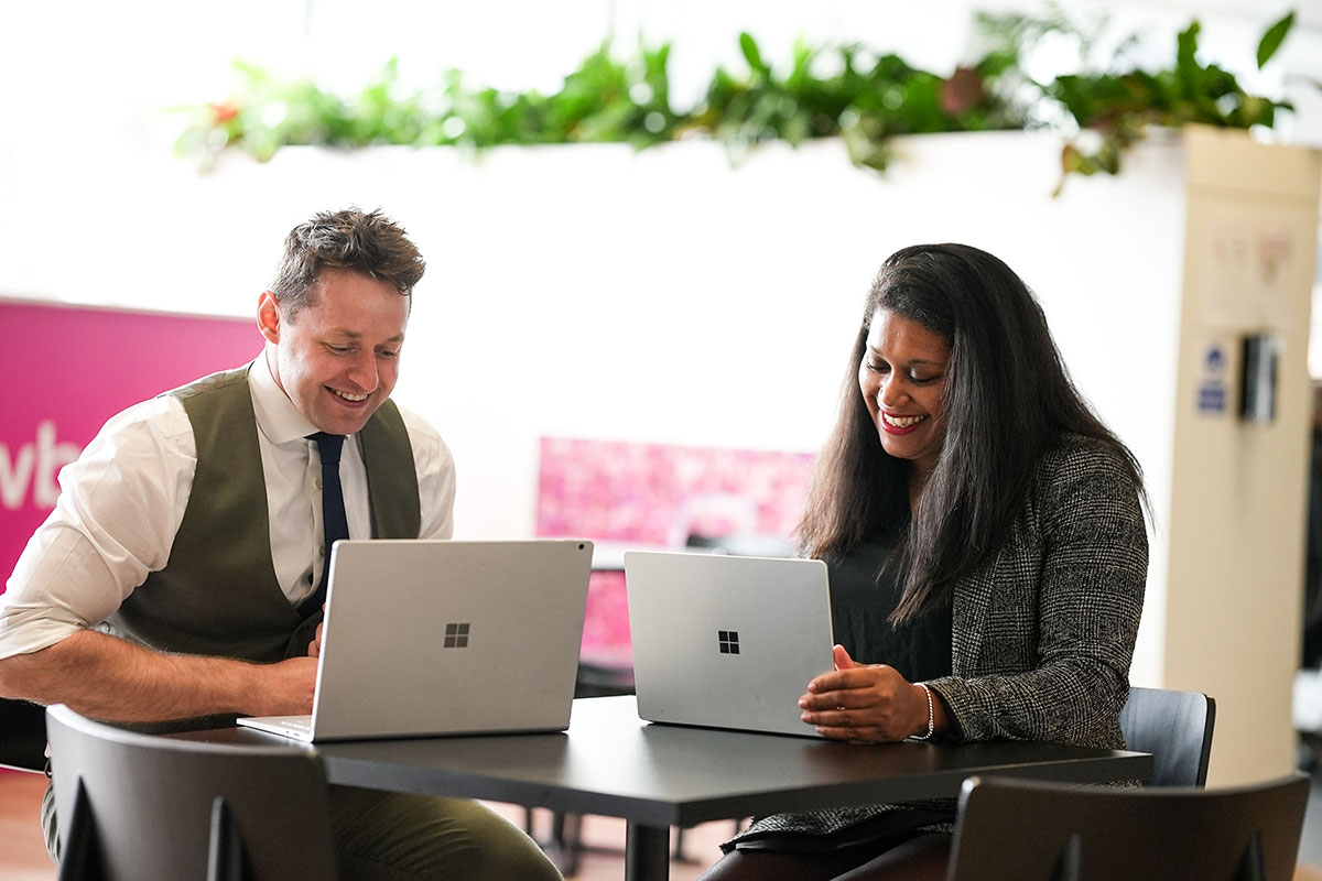 A man and a woman sit at a table, smiling and working on their laptops. They appear to be having a pleasant conversation in a bright, modern office space with plants in the background.