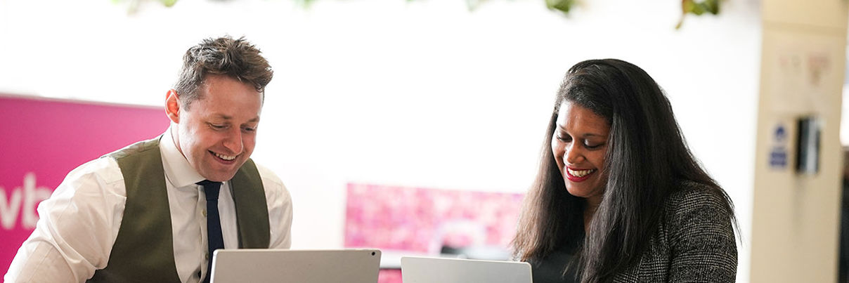 A man and a woman sit at a table, smiling and working on their laptops. They appear to be having a pleasant conversation in a bright, modern office space with plants in the background.