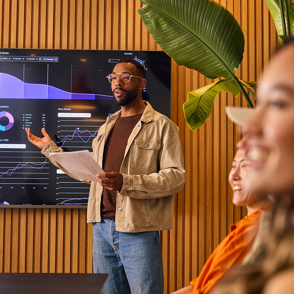 A man holding papers presents data visualizations on a screen to a group seated at a conference table in a modern office with wood paneling and plants.