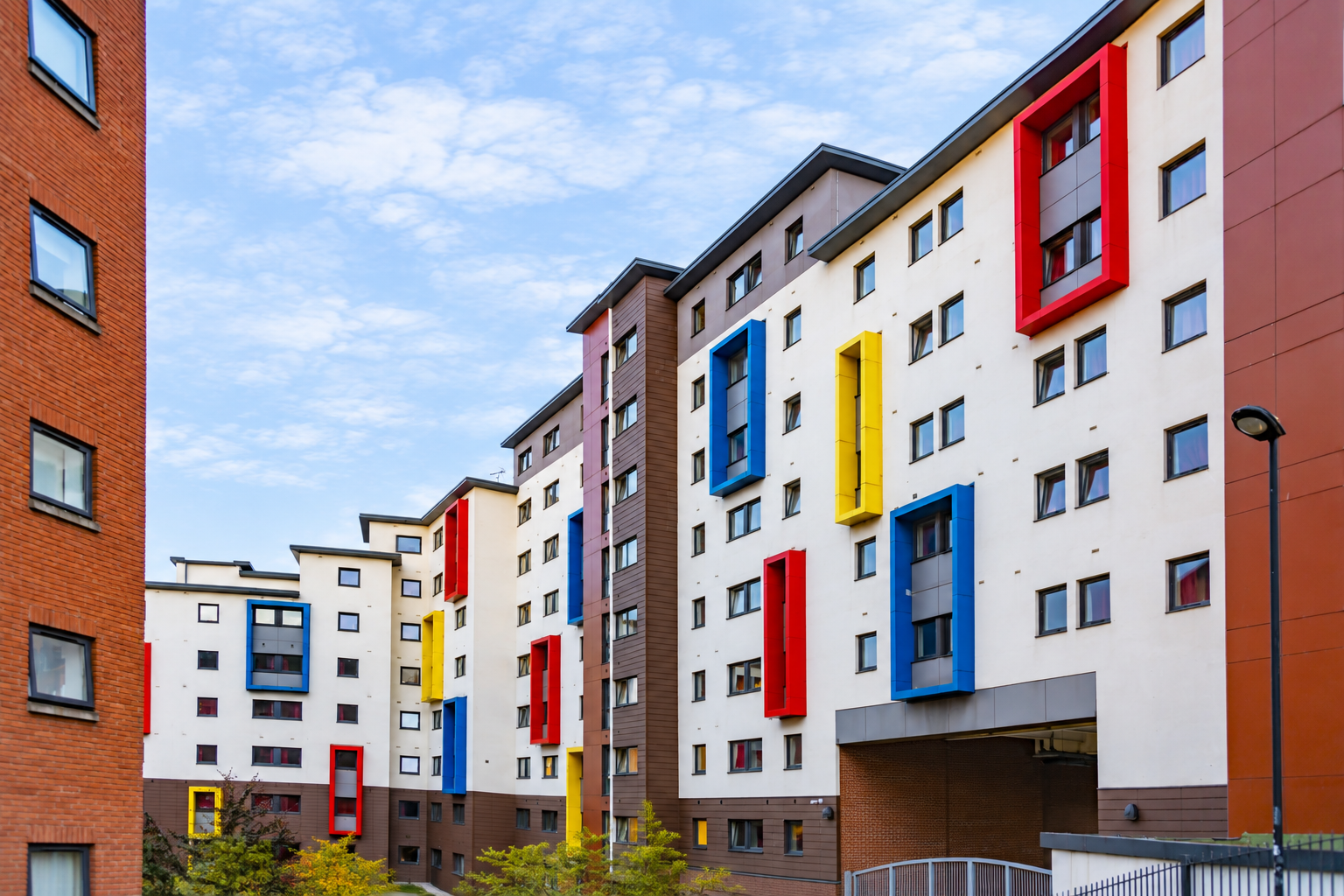 Modern apartment buildings with white facades and brown accents, featuring large, colorful window frames in red, yellow, and blue. The sky is partly cloudy, and there are some trees in the foreground.