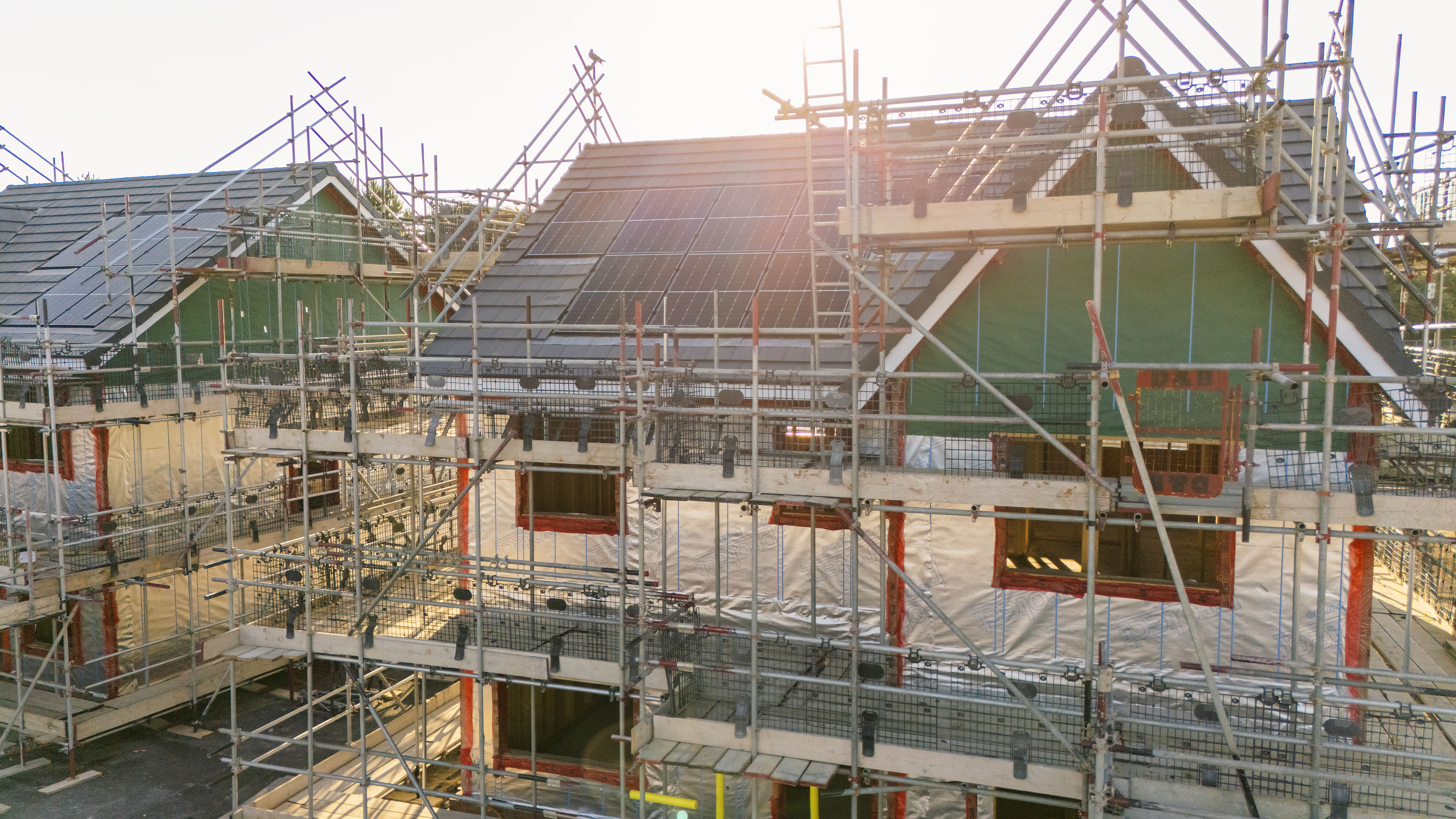 Two houses under construction covered in scaffolding, with unfinished roofs and exposed wooden frames at sunset. Sunlight brightens the image from the background.