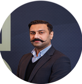 A man with a neatly groomed handlebar mustache, wearing a dark suit jacket, light blue collared shirt, and black sweater, poses in front of a dark blue background with a subtle geometric pattern.