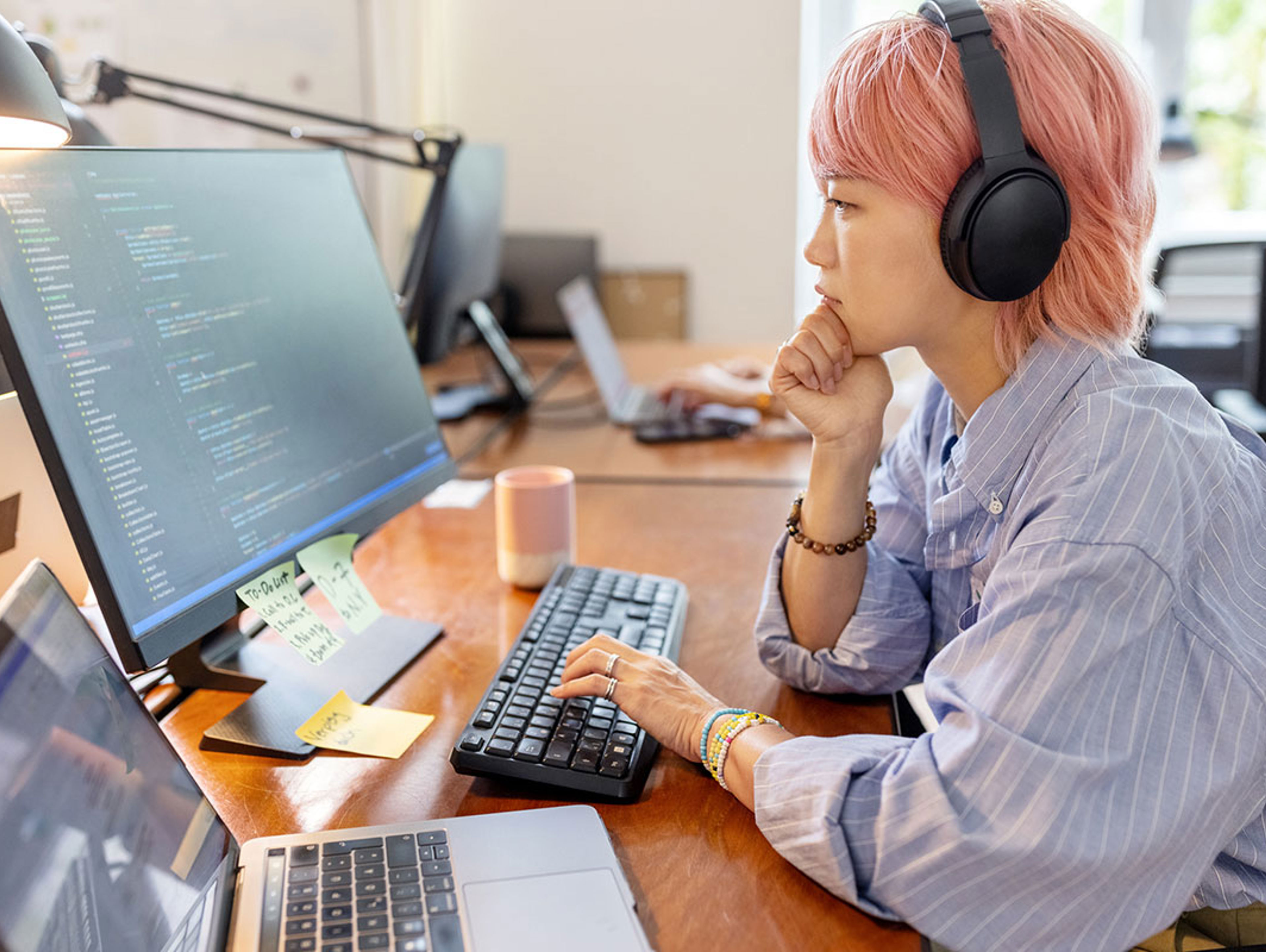 A person with pink hair wearing headphones sits at a desk, focused on a large monitor displaying code. A laptop, keyboard, mouse, and sticky notes are also on the wooden desk.