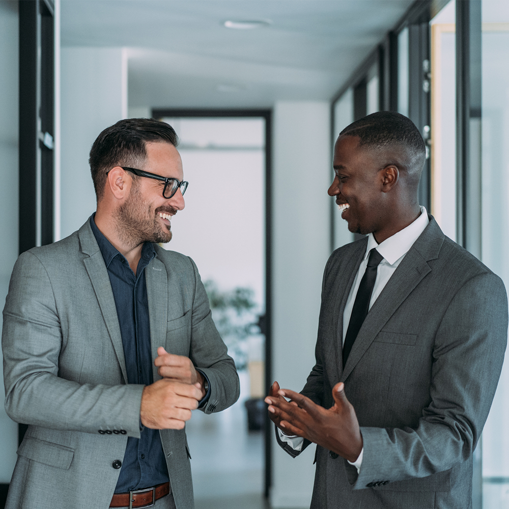 Two professional men in business attire smiling and talking in an office hallway.
