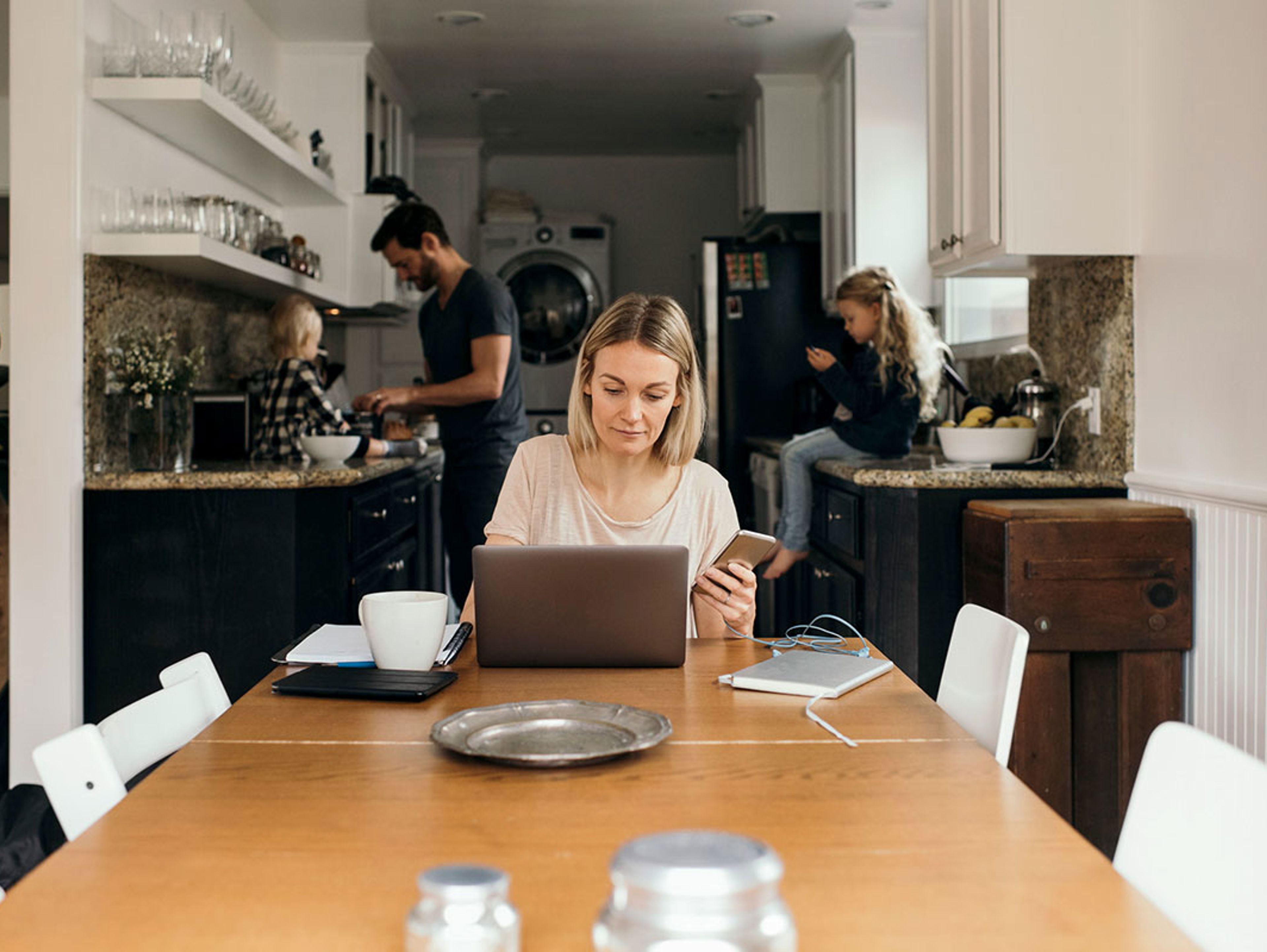 Woman At Kitchen Table Using Mobile With Family