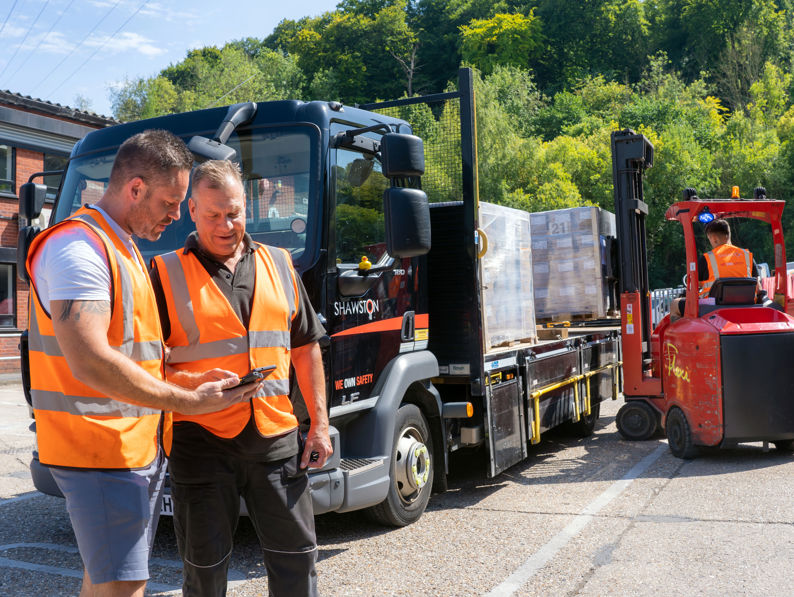 Two men in orange safety vests stand near a truck, looking at a phone. A forklift driver moves a pallet in the background. Trees and a building are visible on a sunny day.