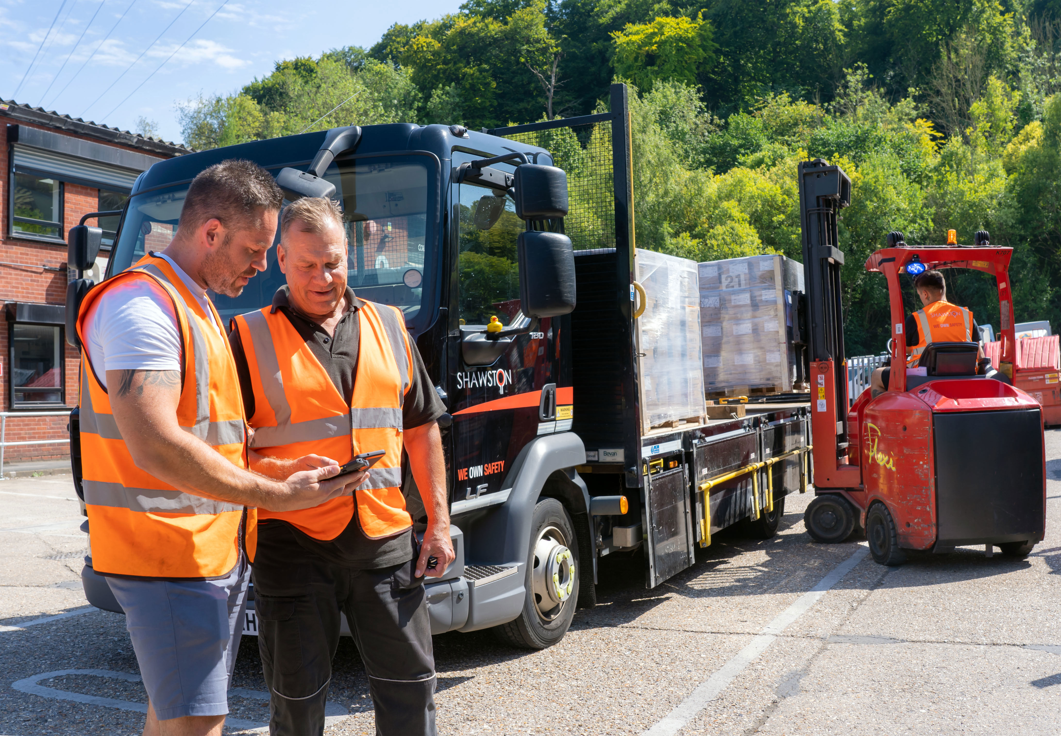 Two men in orange safety vests stand near a truck, looking at a phone. A forklift driver moves a pallet in the background. Trees and a building are visible on a sunny day.