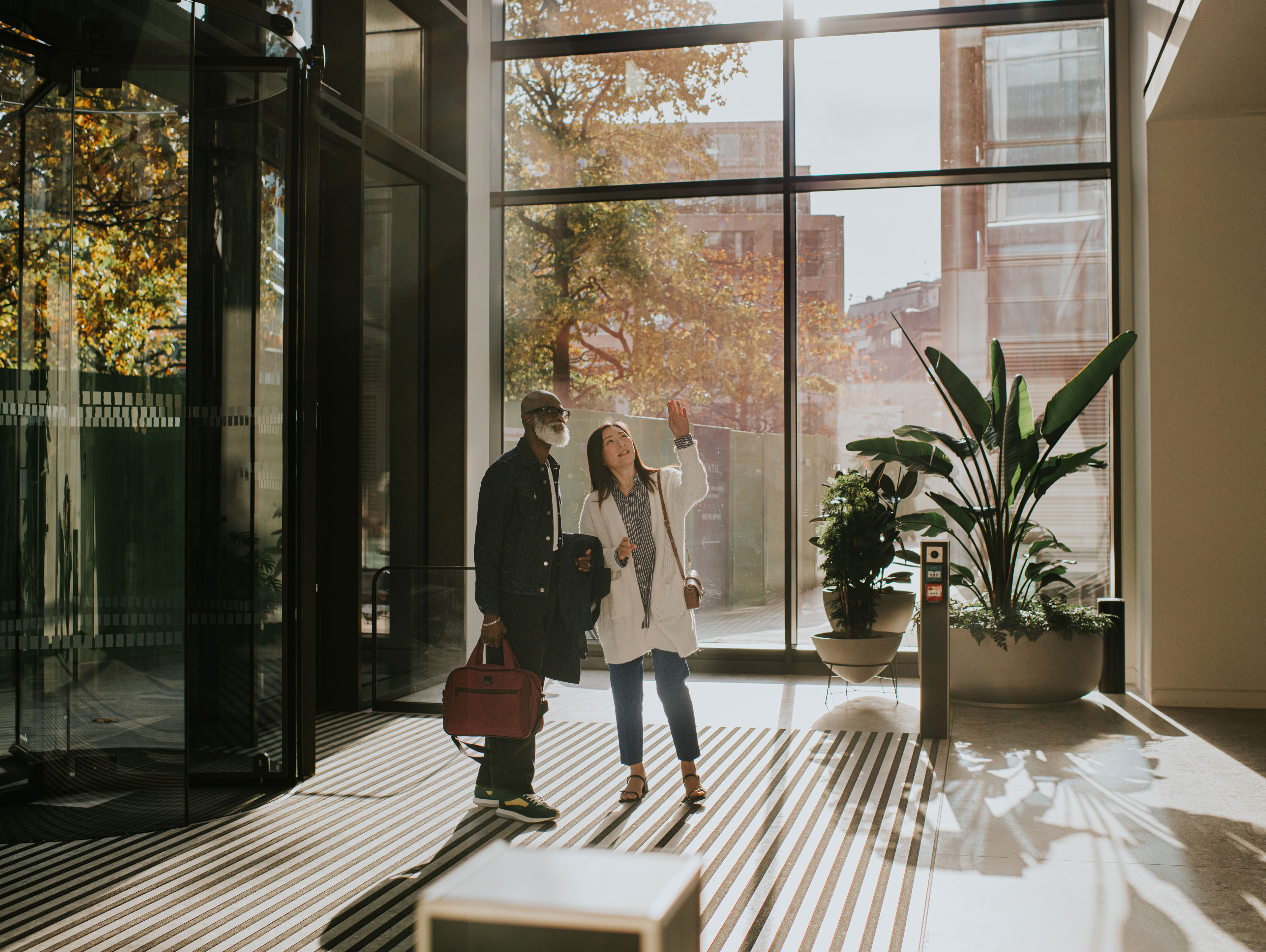 Two people stand in a sunlit, modern building lobby with large windows, one carrying a red bag while the other gestures upward, surrounded by greenery and shadows cast on the floor.